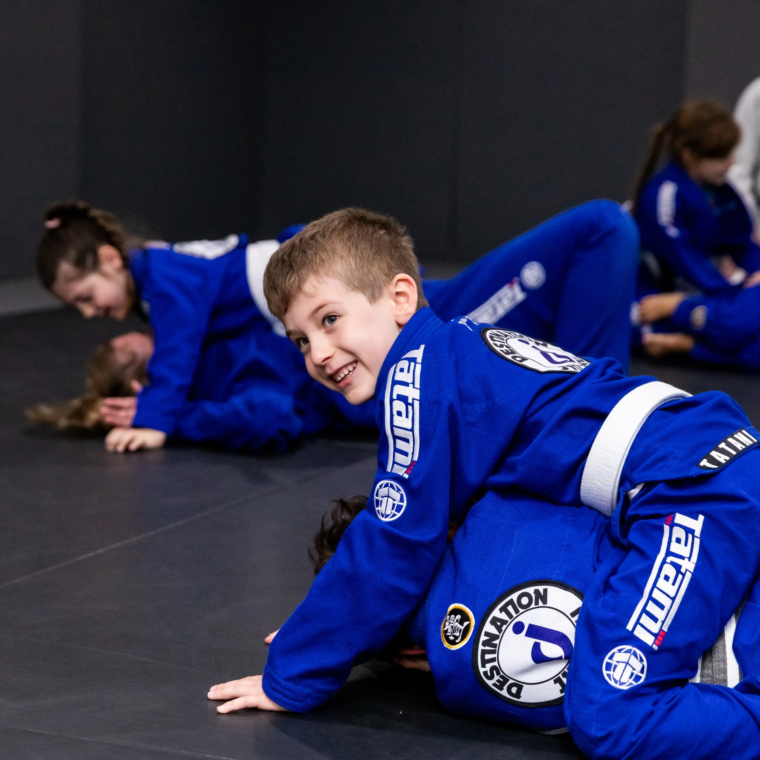A class with 6 children training on the floor. A young boy with blonde hair on top of another young boy with dark brown hair. All the children are wearing bright blue gis. 