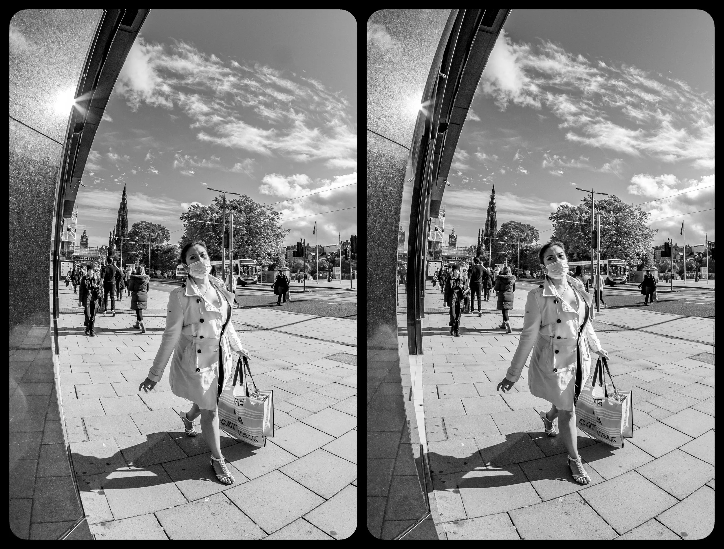 A woman wearing a face mask, trench coat, and heels walking with shopping bags outside near a reflective wall, with pedestrians, trees, and a church spire in the background, on a sunny day.