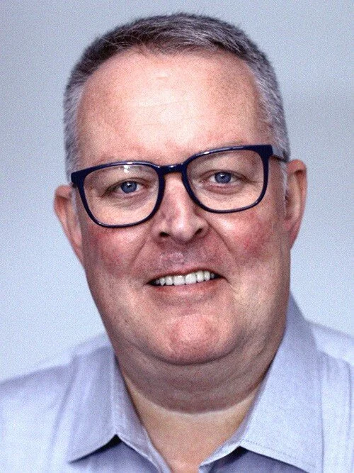 A man with glasses smiling, wearing a light shirt against a neutral background.