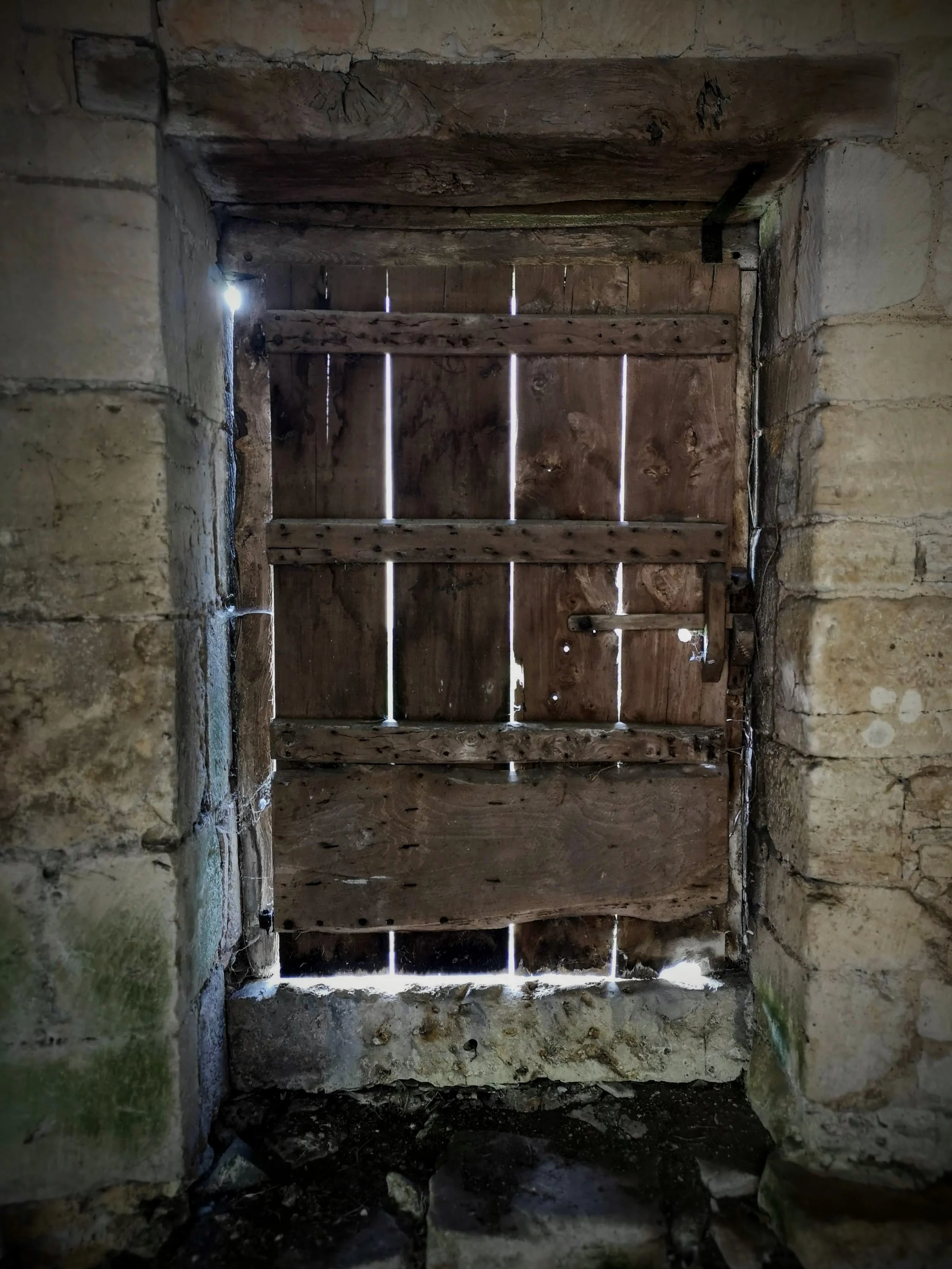 Old wooden door with gaps between the slats, set in a stone wall, with light shining through the gaps.