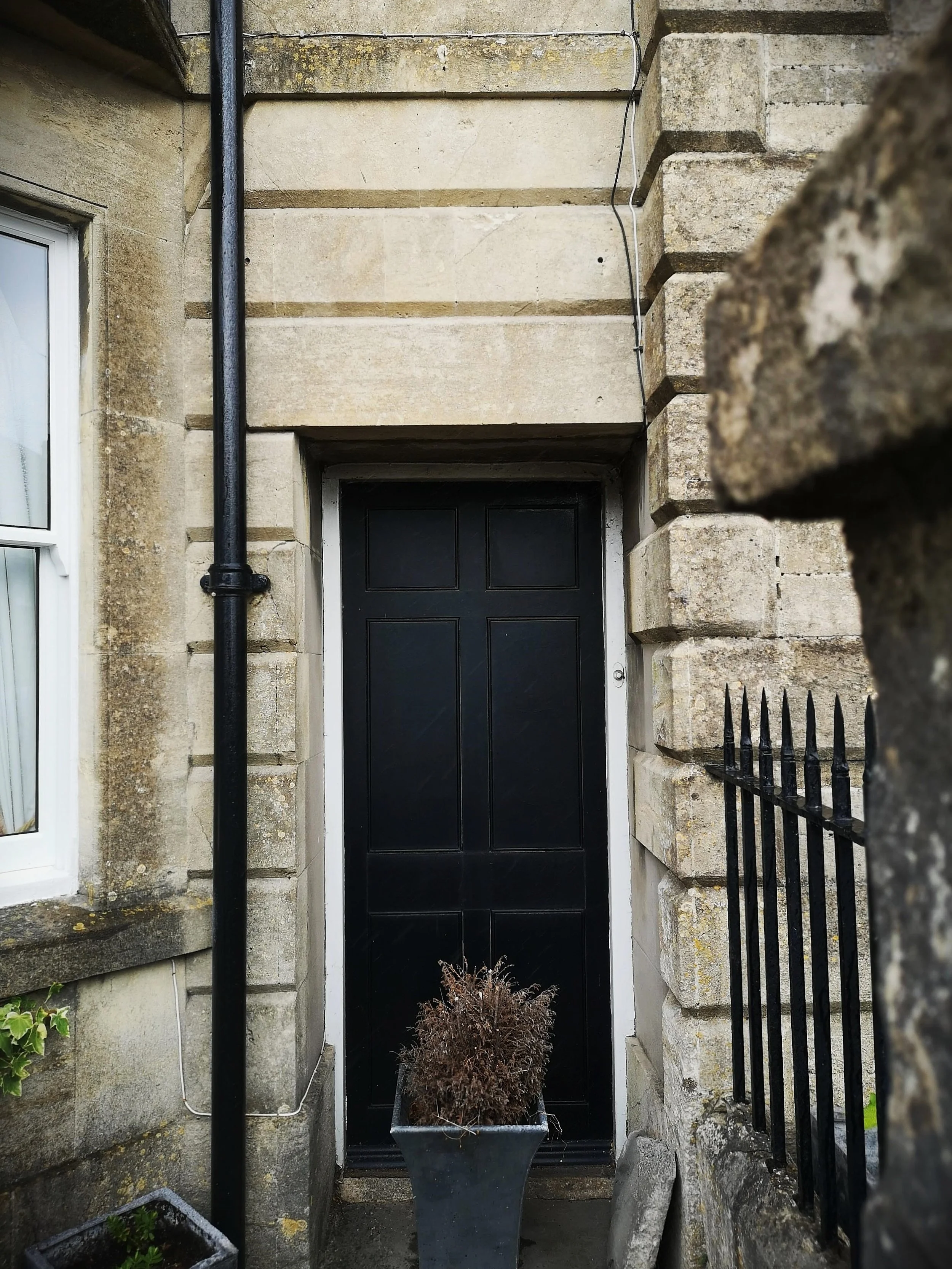 Black door framed by stone walls, with a plant in a gray pot in front and a black iron fence to the right.