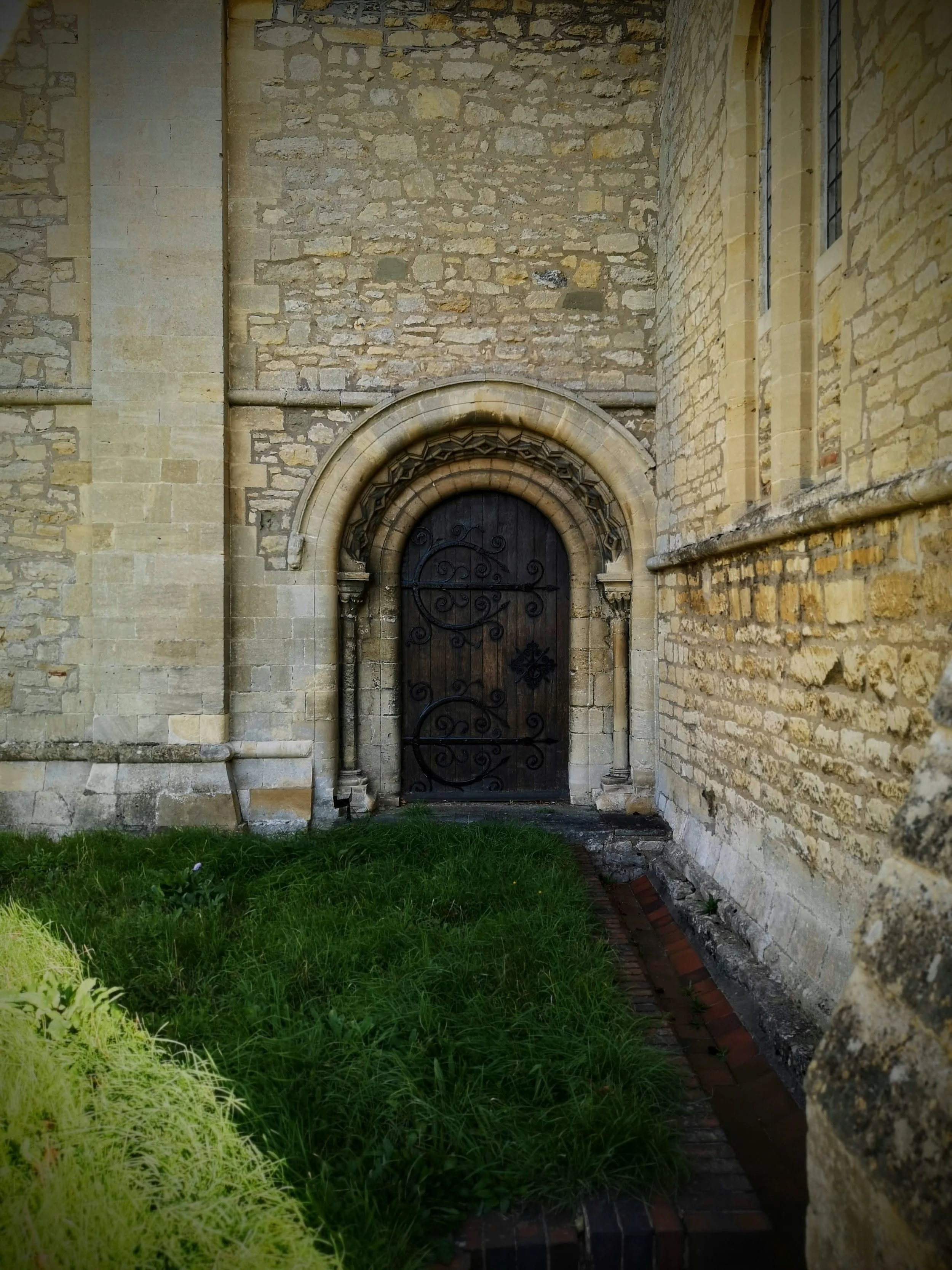 Old stone church with a black wooden door surrounded by an ornate stone archway, and narrow grassy area in front.
