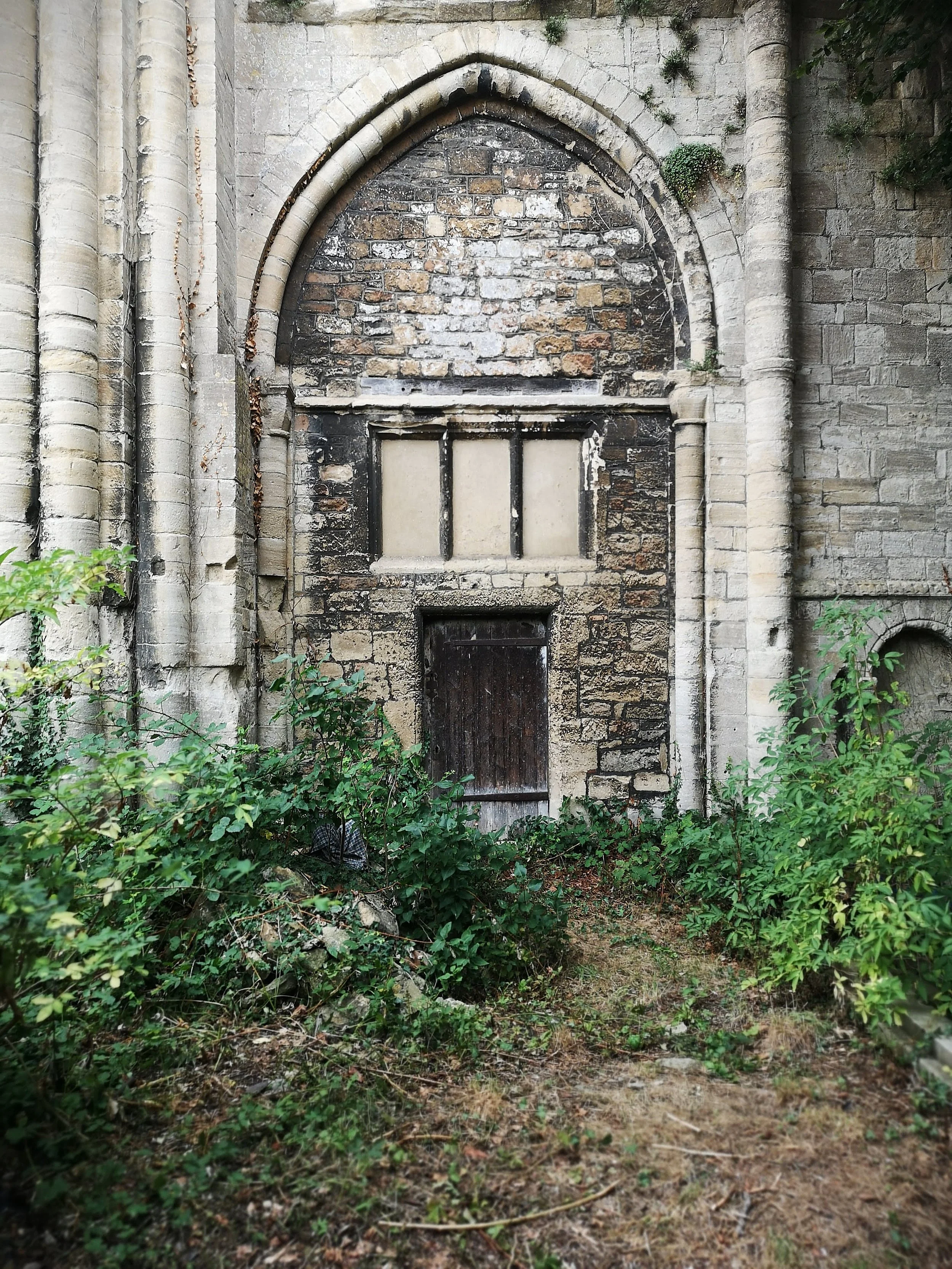 Old stone building facade with a closed, weathered door and a large arched window above, surrounded by overgrown bushes.