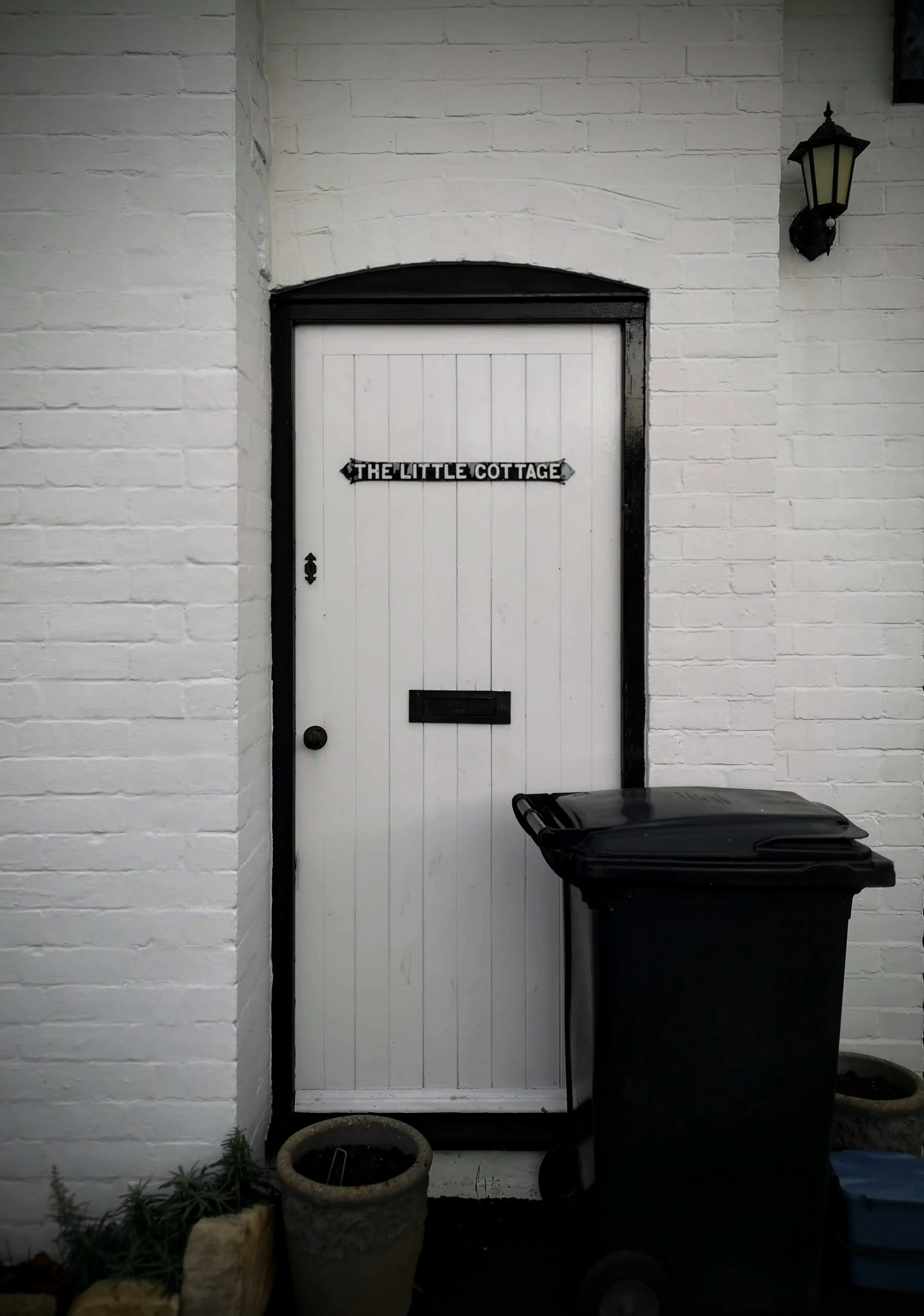 A white door with black trim on a white brick wall, labeled "The Little Cottage," with a black trash bin and a flower pot nearby.