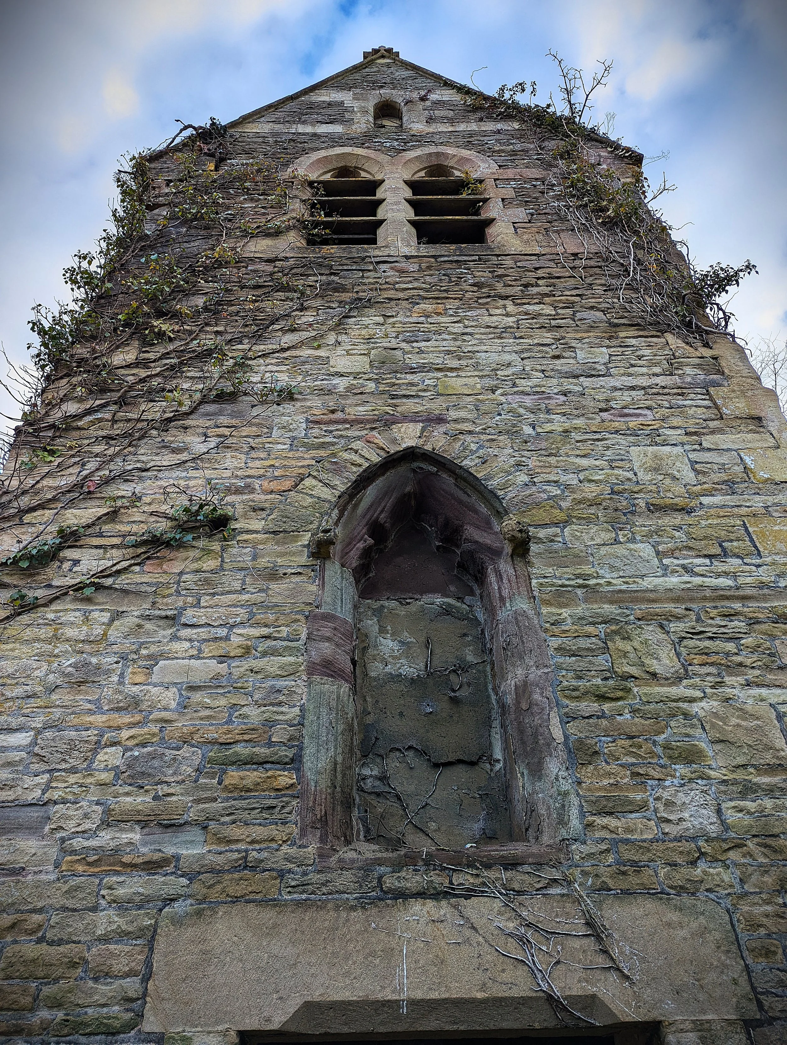 An old, abandoned stone church tower with vines growing on the walls, arched window openings near the top, and a weathered, arched window or doorway at the bottom against a cloudy sky.
