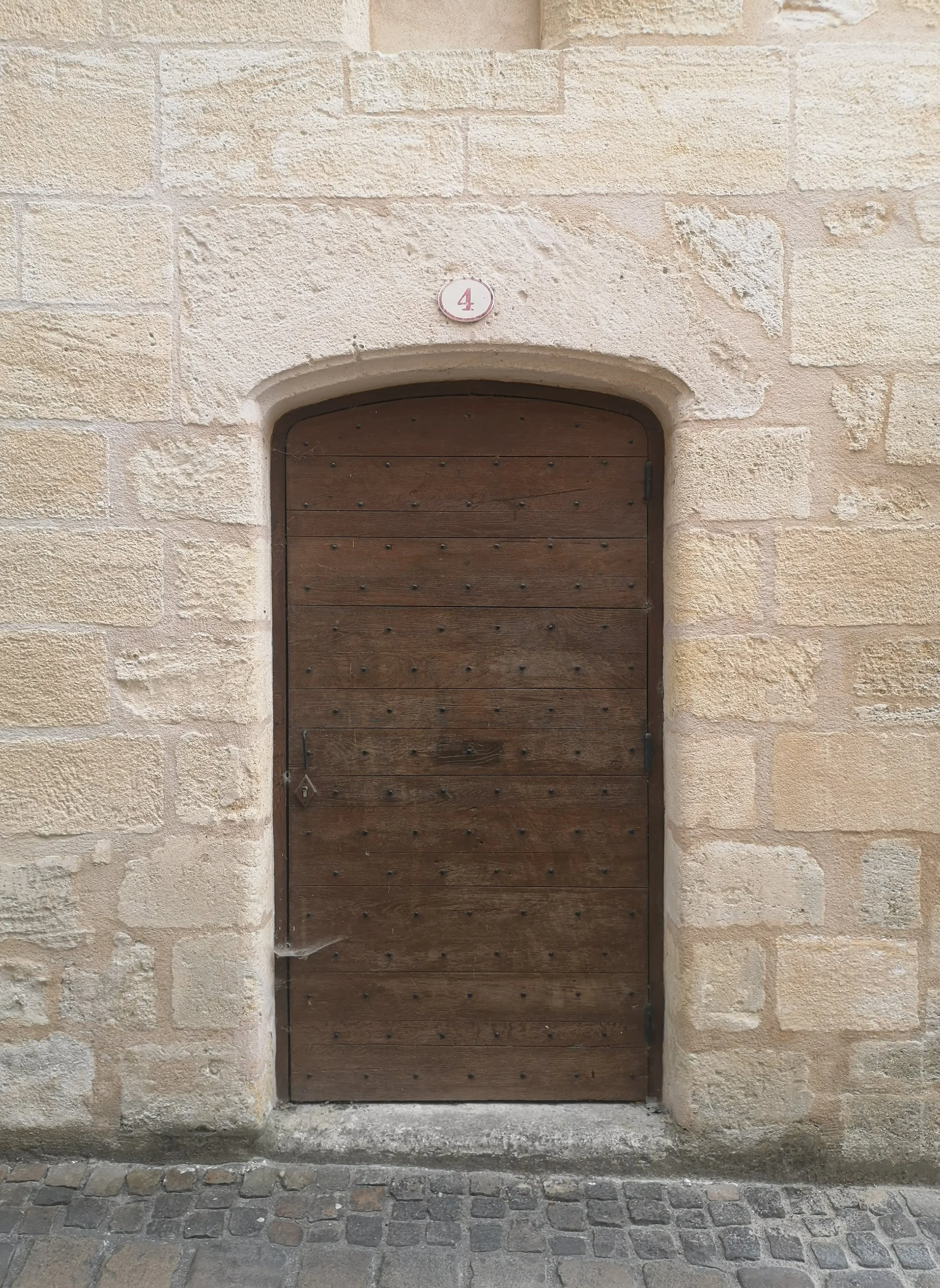 A small arched wooden door set into a beige stone wall, with a small round plaque above it displaying the number 4.