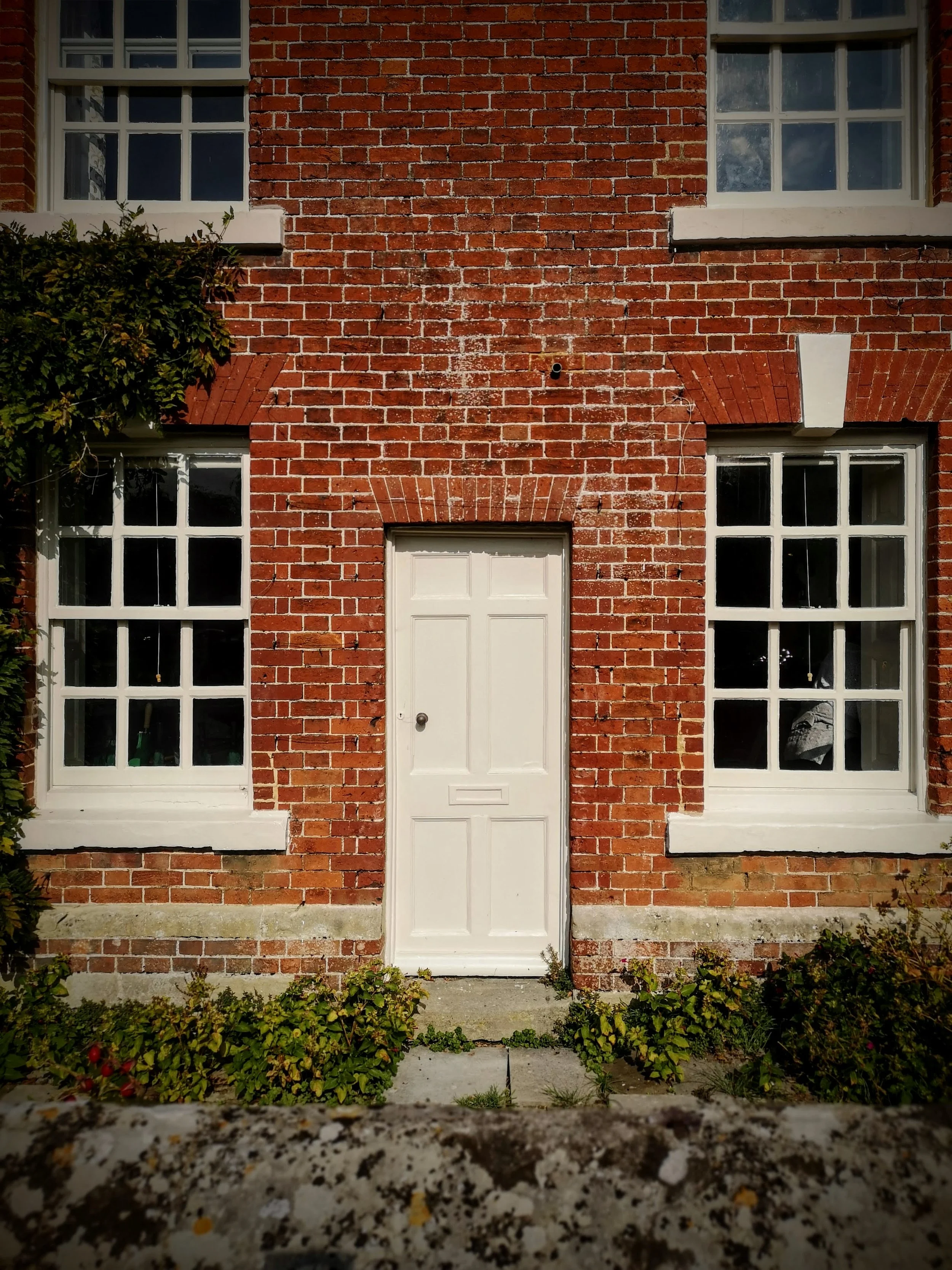 Front of brick house with white door and large white-framed windows on either side. Small garden with plants and flowers at the base of the house.