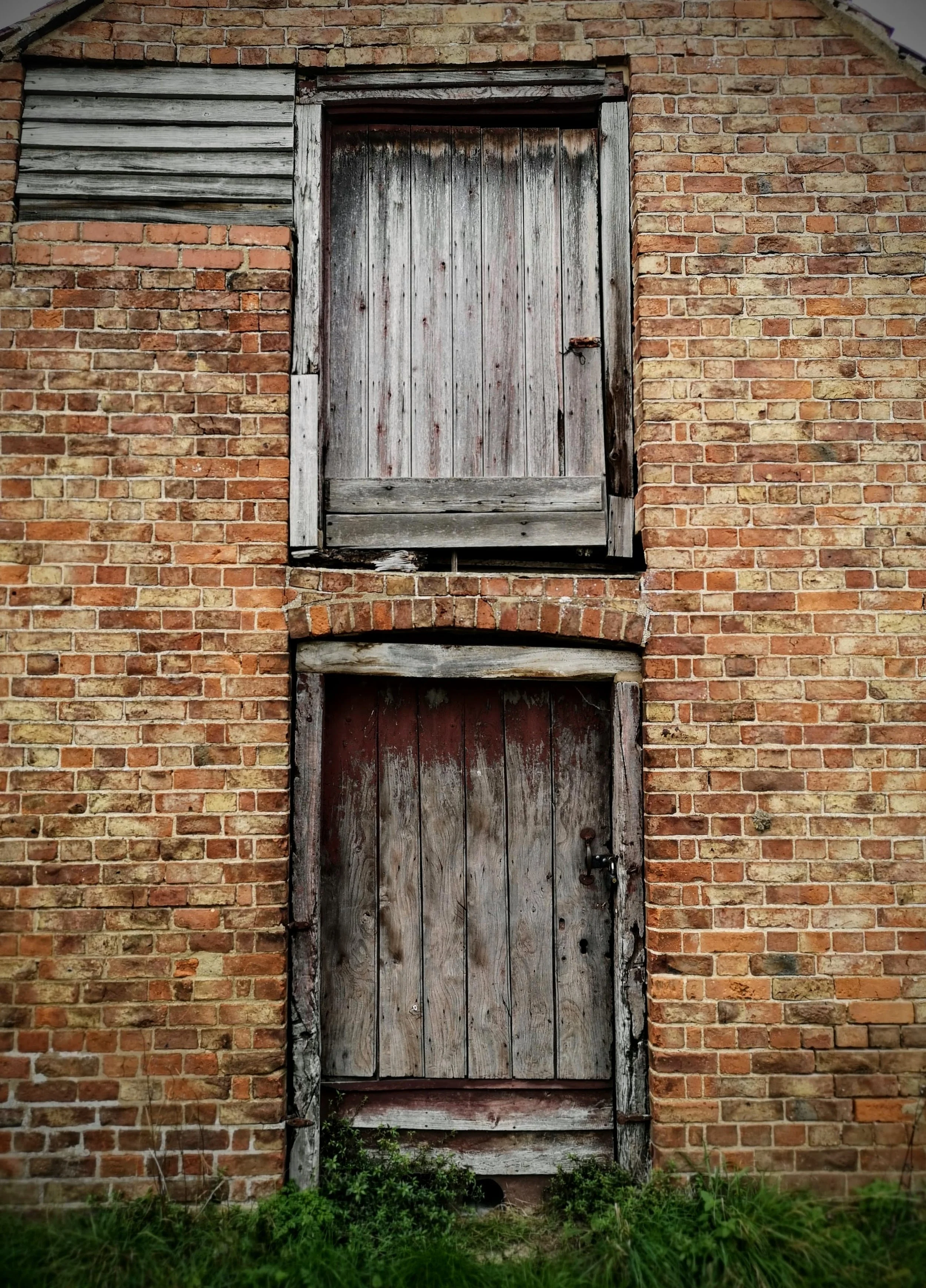 An old brick building with two weathered wooden doors, one above the other, both with chipped paint and rusty hardware, surrounded by green grass and weeds.