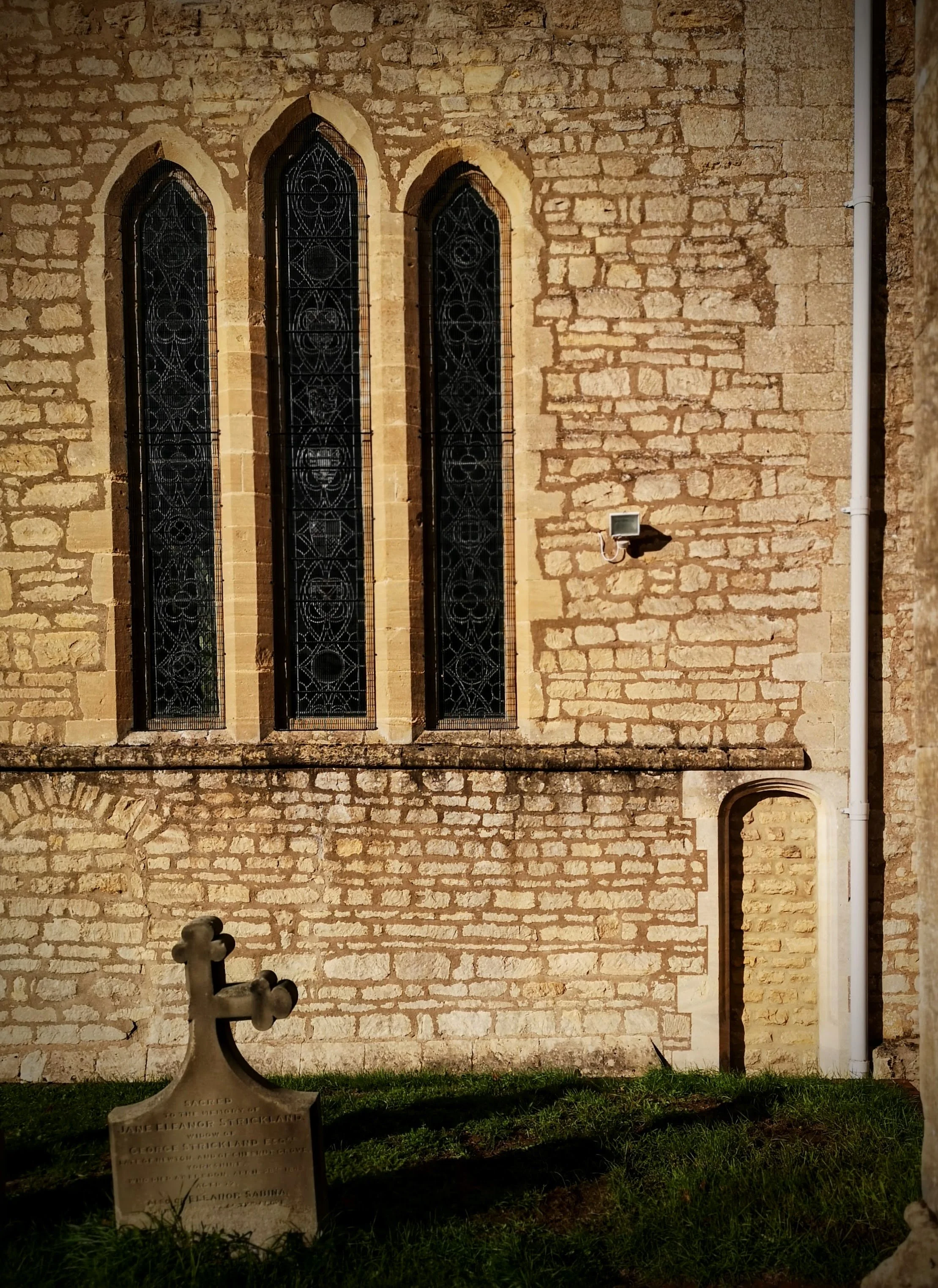 A stone church wall with three tall, narrow Gothic-style windows with black decorative stained glass. In front, there's a small grave marker shaped like an anchor for Jane Eleanor Stickland, who was the wife of George Stickland, located on a grassy a