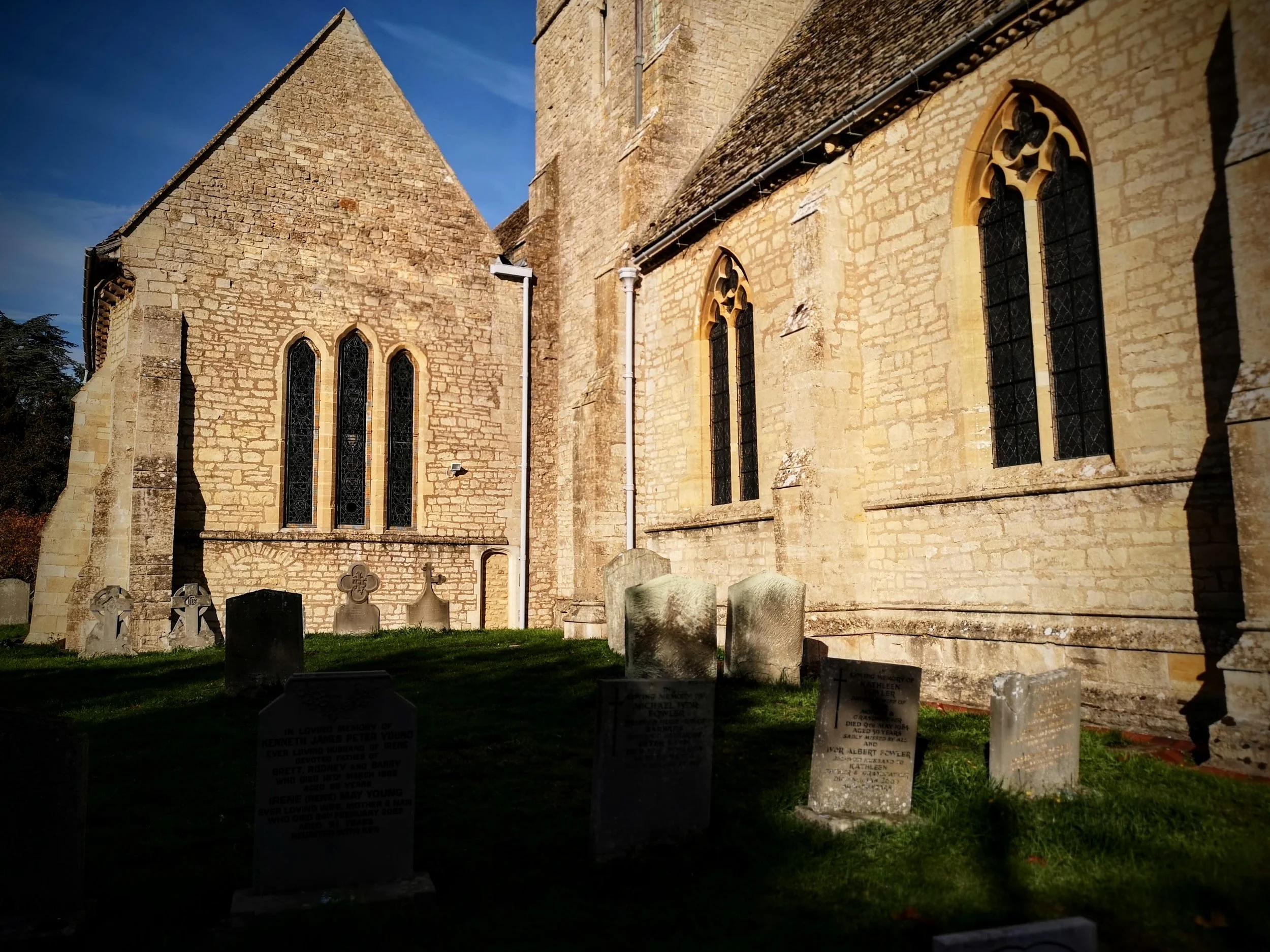 A historic stone church with gothic windows and a small graveyard in front, bathed in late afternoon sunlight.