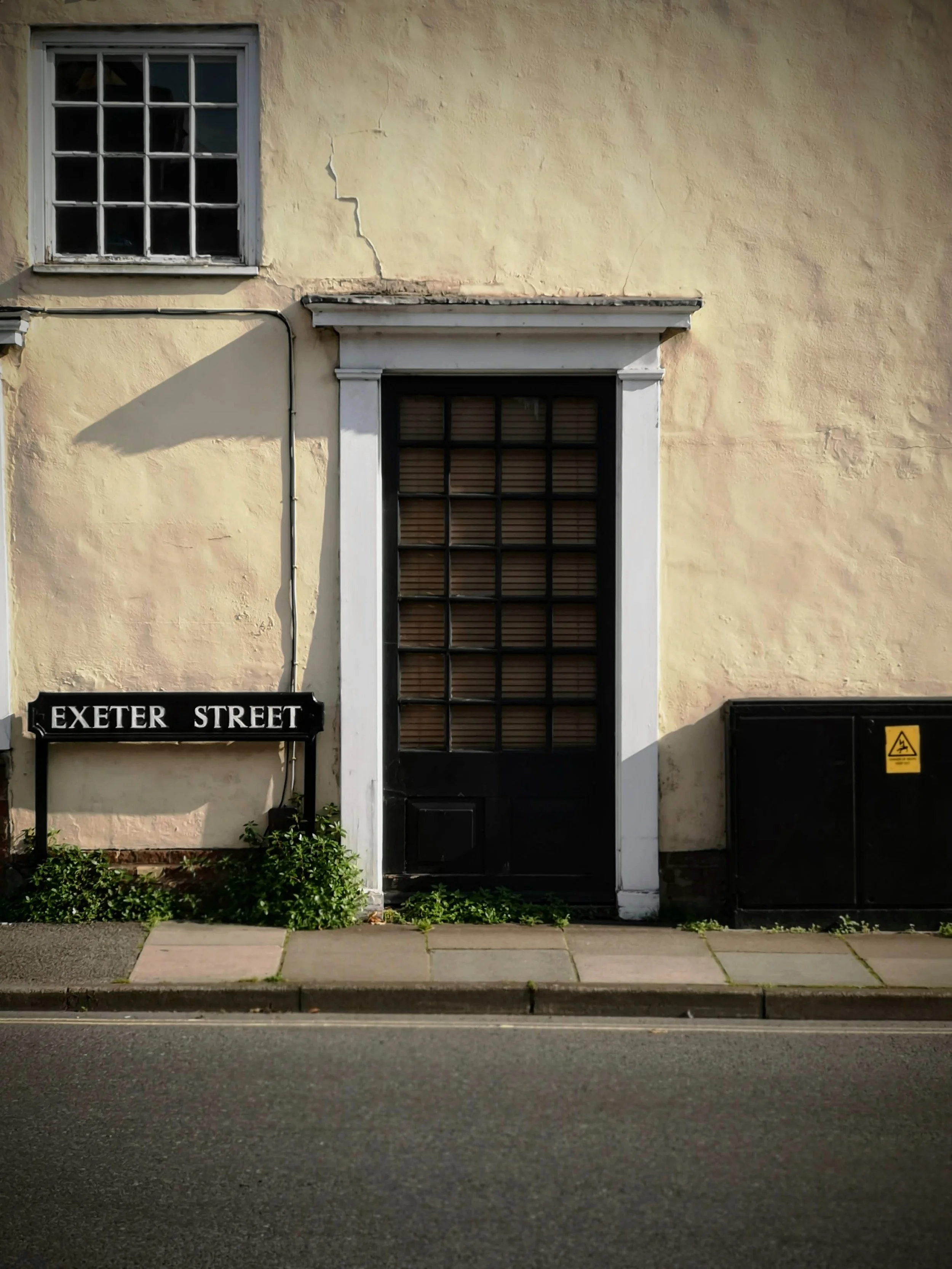 A beige building with a black door and window, a street sign that reads 'Exeter Street', some green plants, and a utility box.