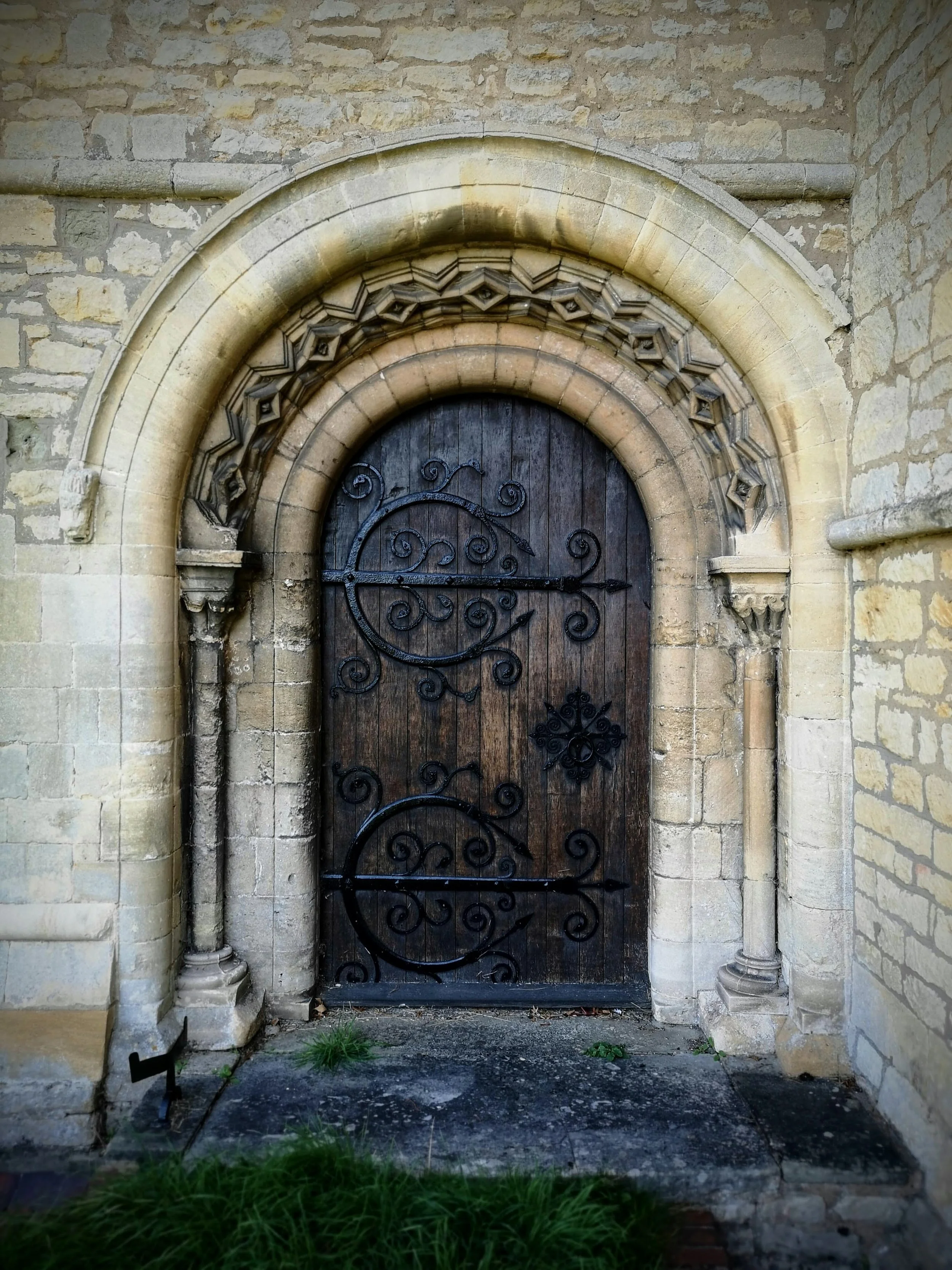 An arched wooden door with intricate black wrought iron decorations, set in a stone wall with detailed stone carvings around the arch.
