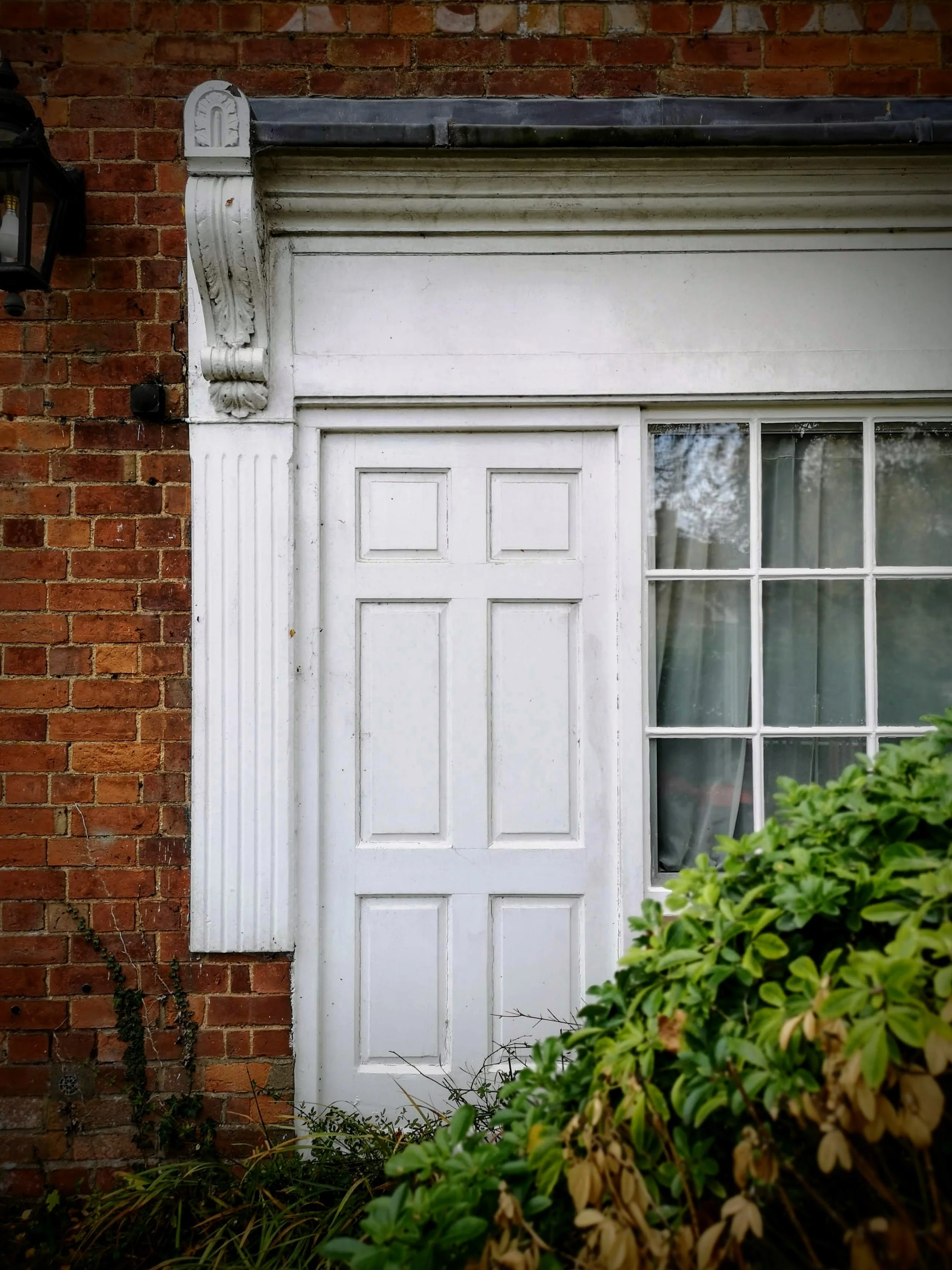 White wooden door with a grid window next to a brick wall, partially obscured by green plants, with decorative white trim and an exterior lantern light.