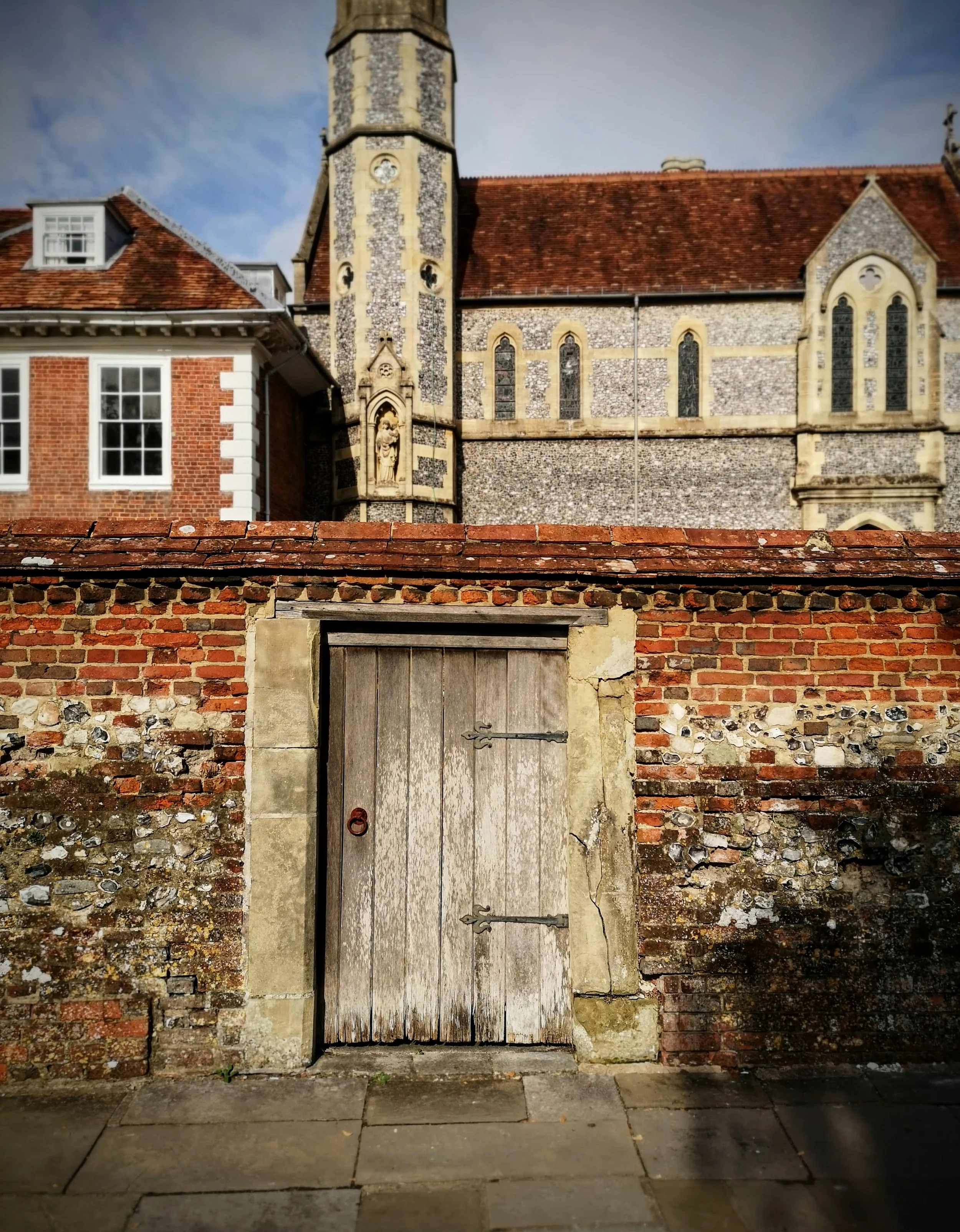 An old wooden gate with metal hinges set into a brick and stone wall in front of a historic church with stone walls, arched windows, and a tall tower with decorative stonework.