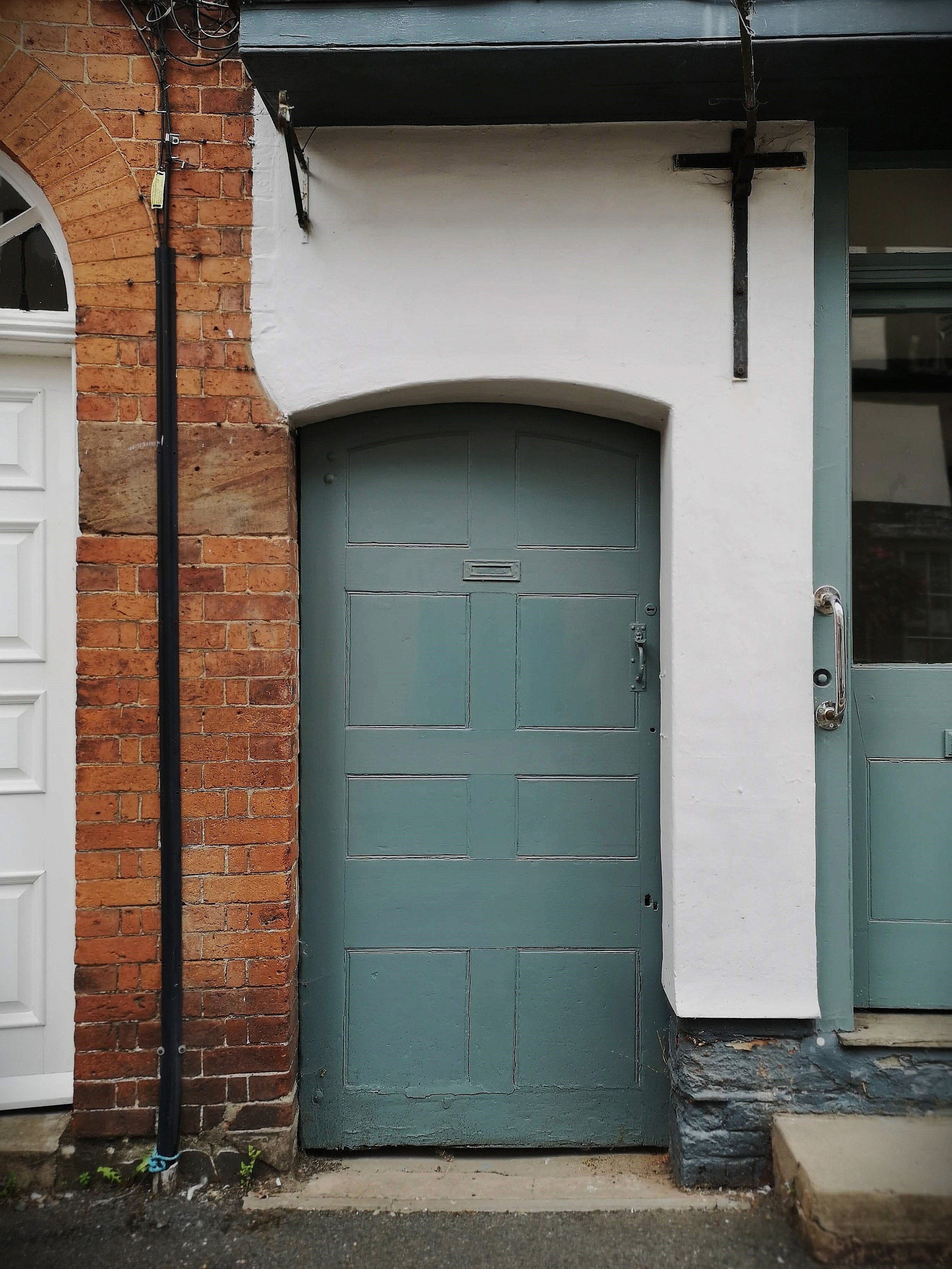 A teal-colored door with a horizontal mail slot, set within a white wall. To the left is a brick wall with an arched window, and to the right is a glass-paneled door with a metal handle. Overhead, part of a black metal awning and support are visible.