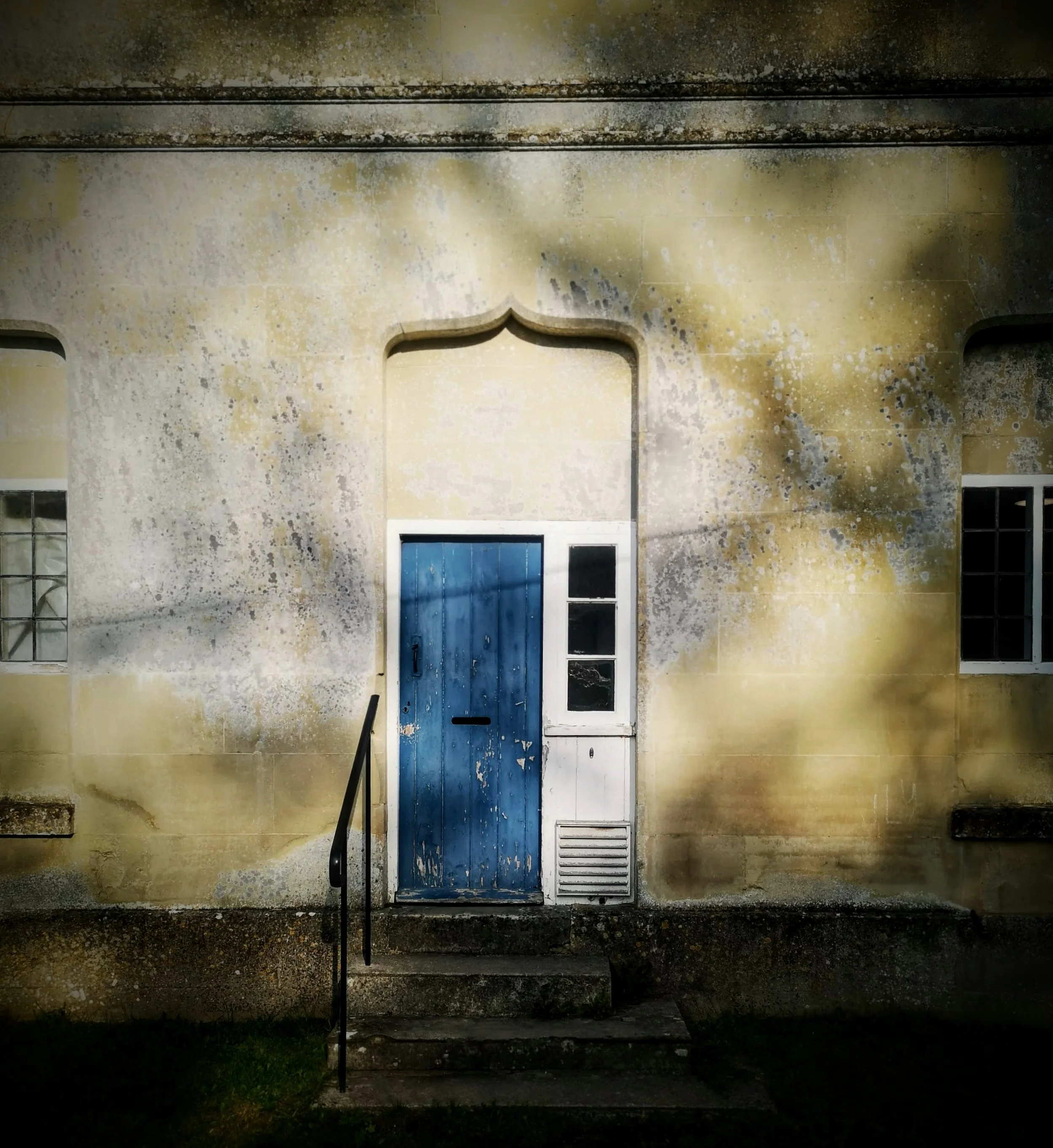 Old yellow building with peeling paint, blue door, small set of stairs, and black handrail, shadows cast on the wall.