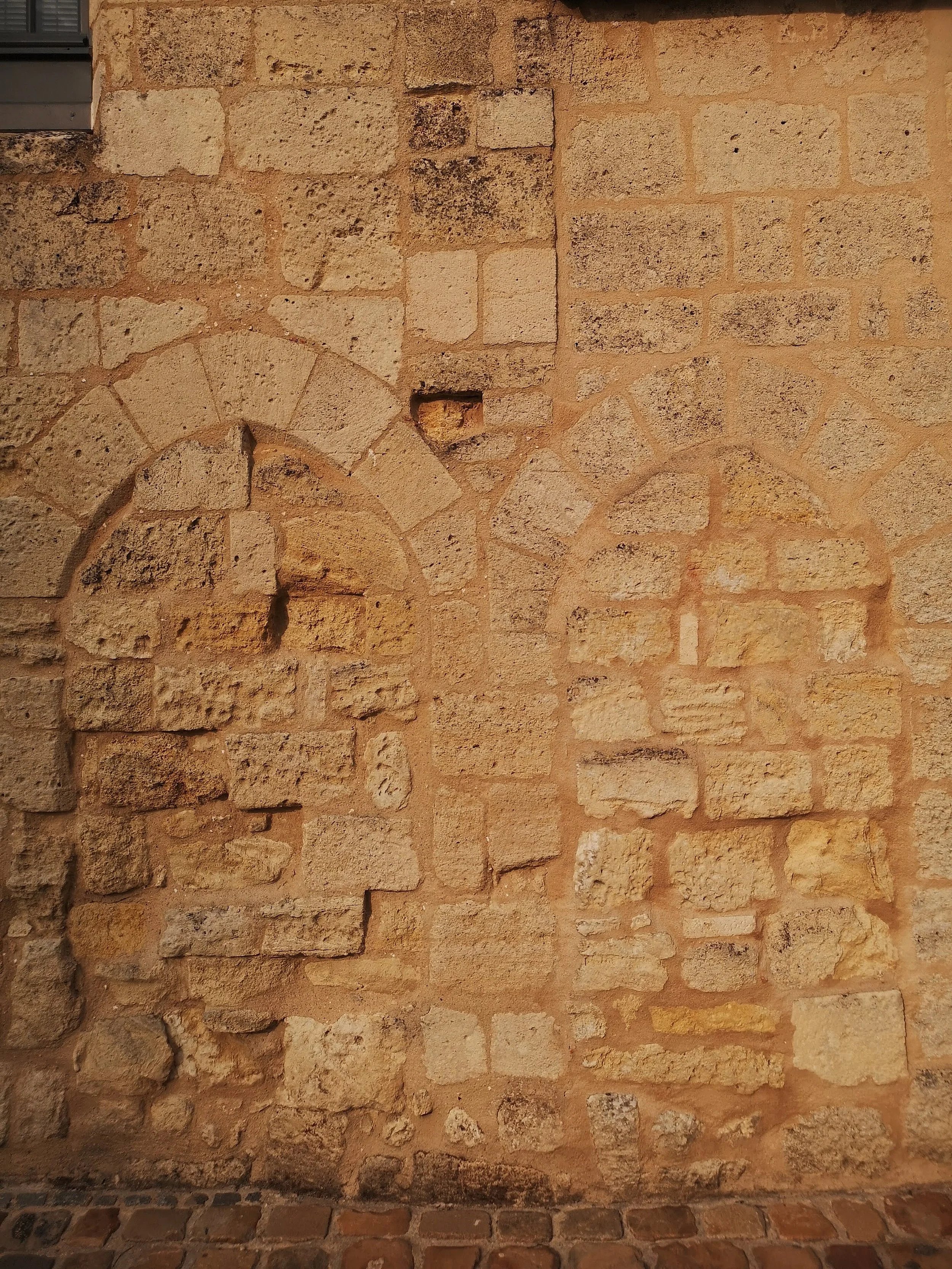 A close-up of an old stone wall with two arch-shaped indentations, one missing bricks, and a small square hole