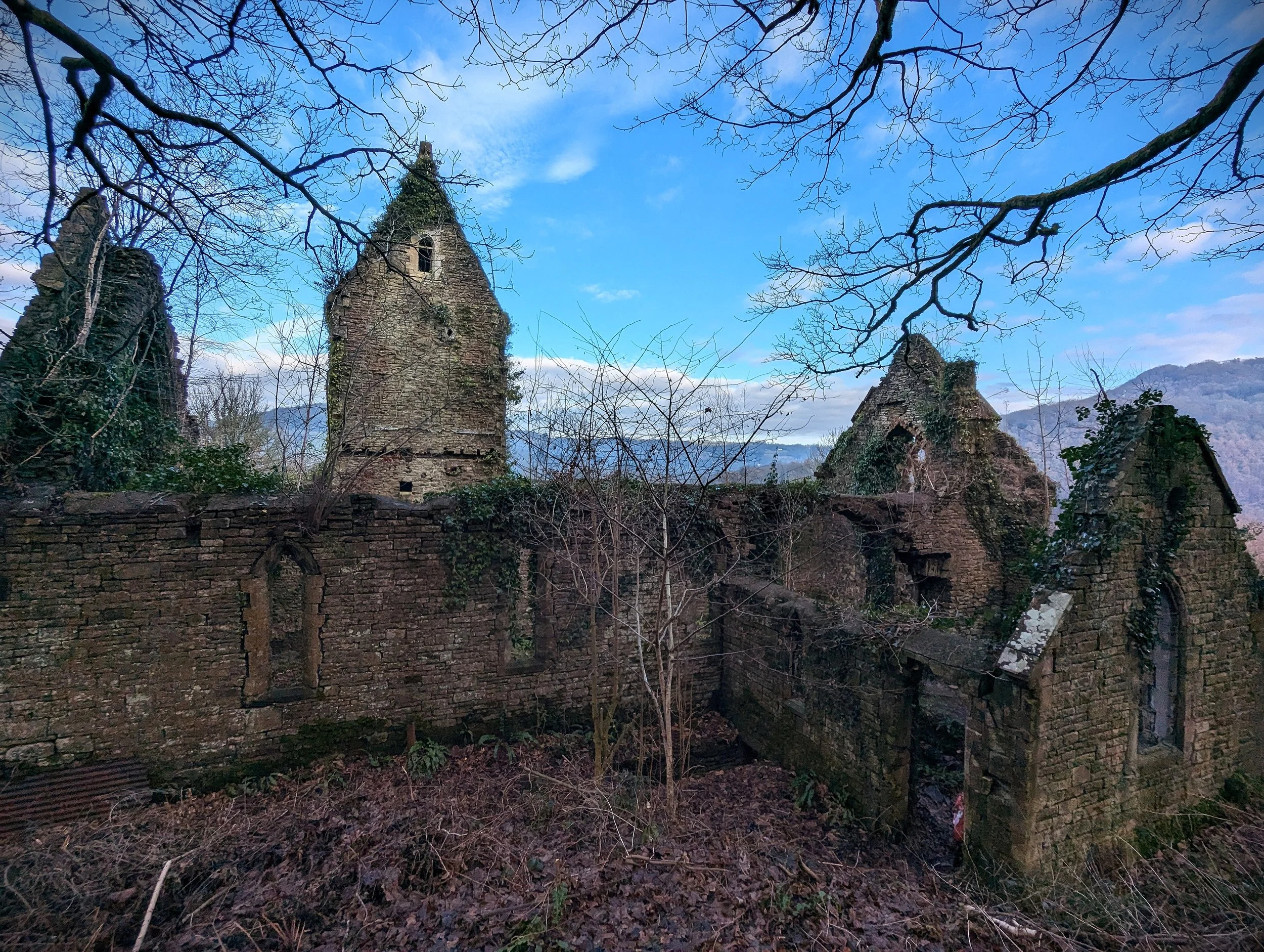 Ruins of an old stone building with arched windows, surrounded by leafless trees and mountains in the background under a blue sky.