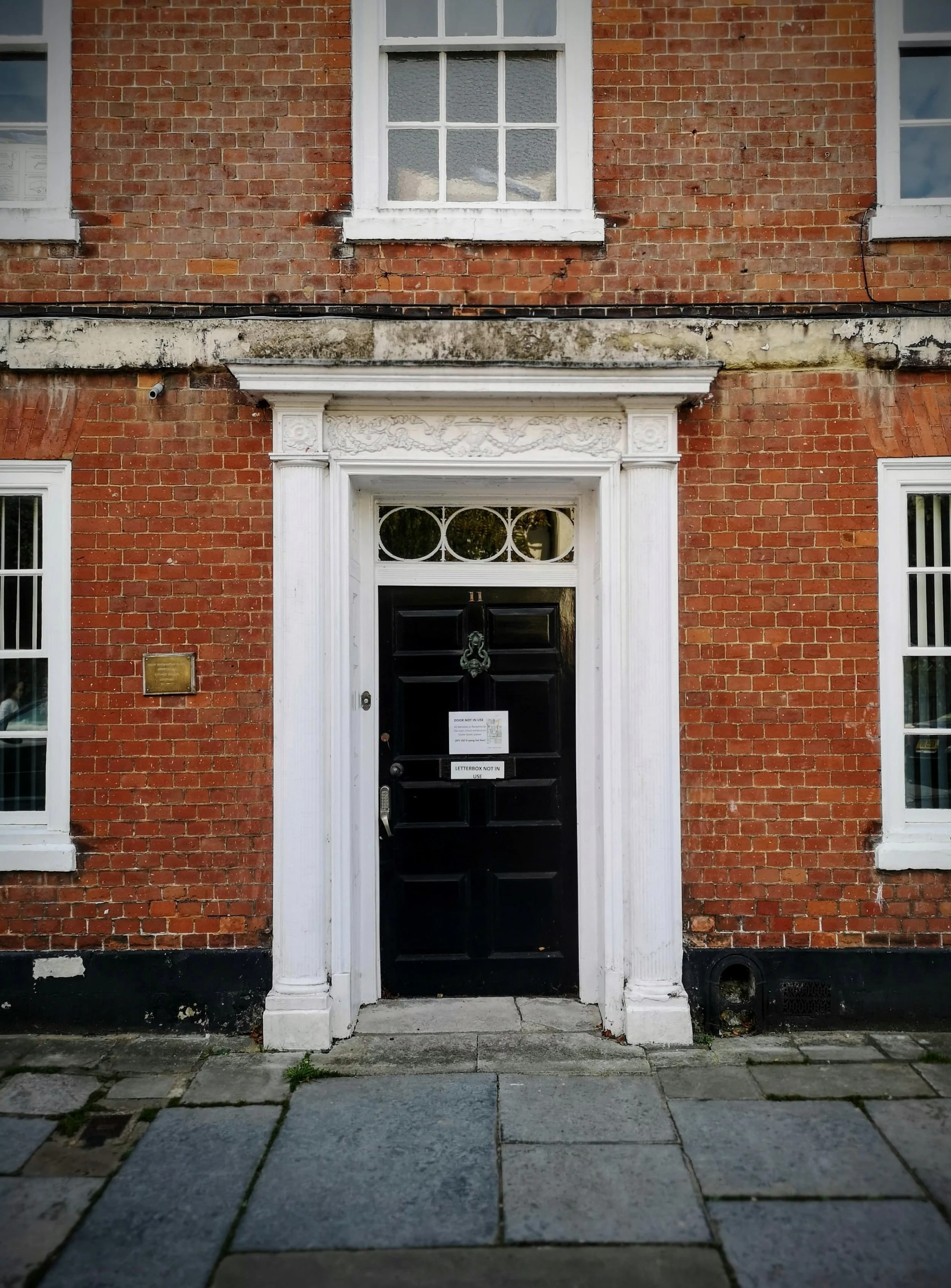 Black front door with decorative white trim on a red brick building. Small signs attached to the door. Two windows above the door and two more on the sides, all with white window frames.