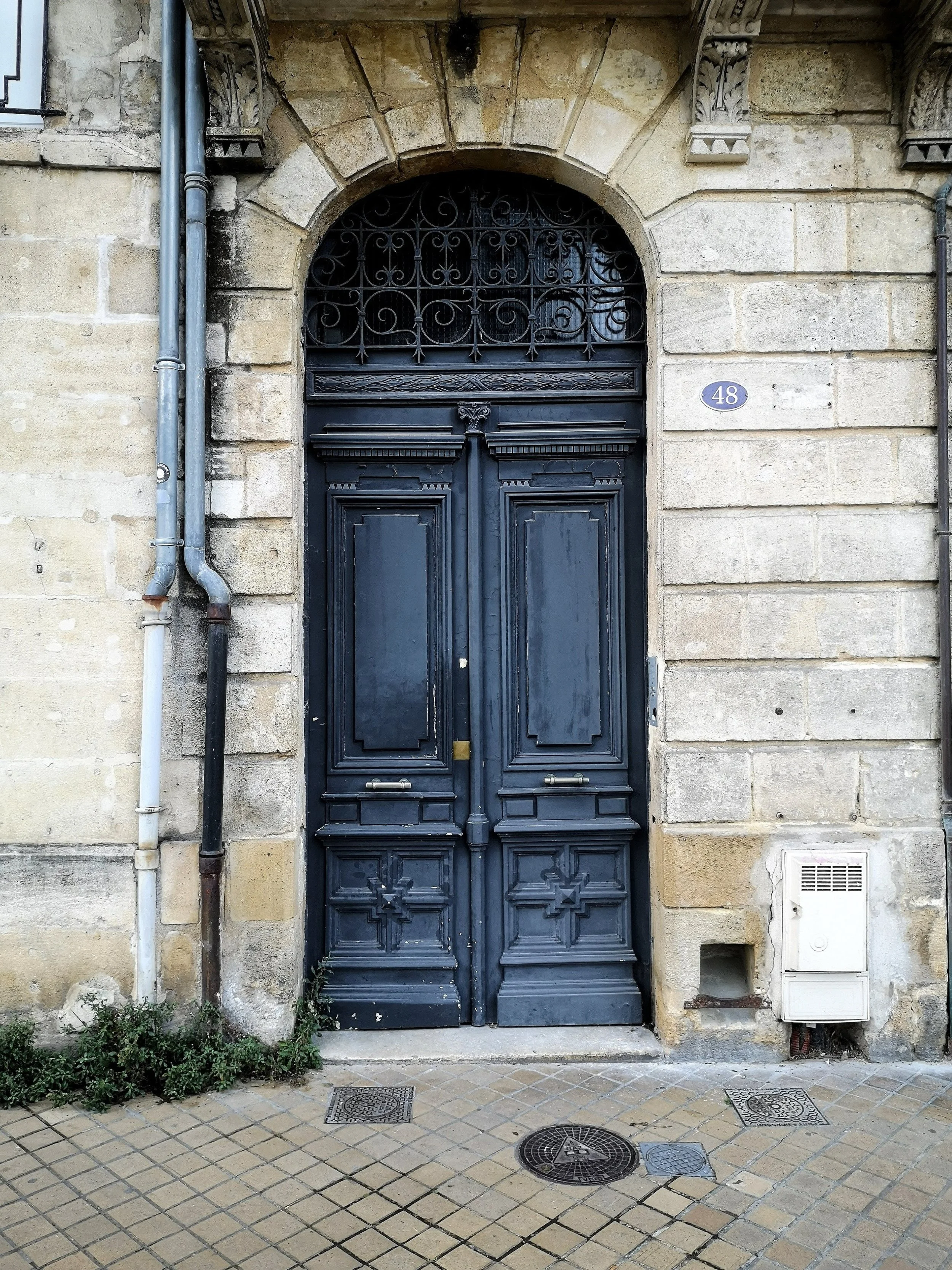 A vintage black wooden door with decorative panels and an arched wrought iron grille window above, set in a stone building with a street number 48 on a small plaque to the right.