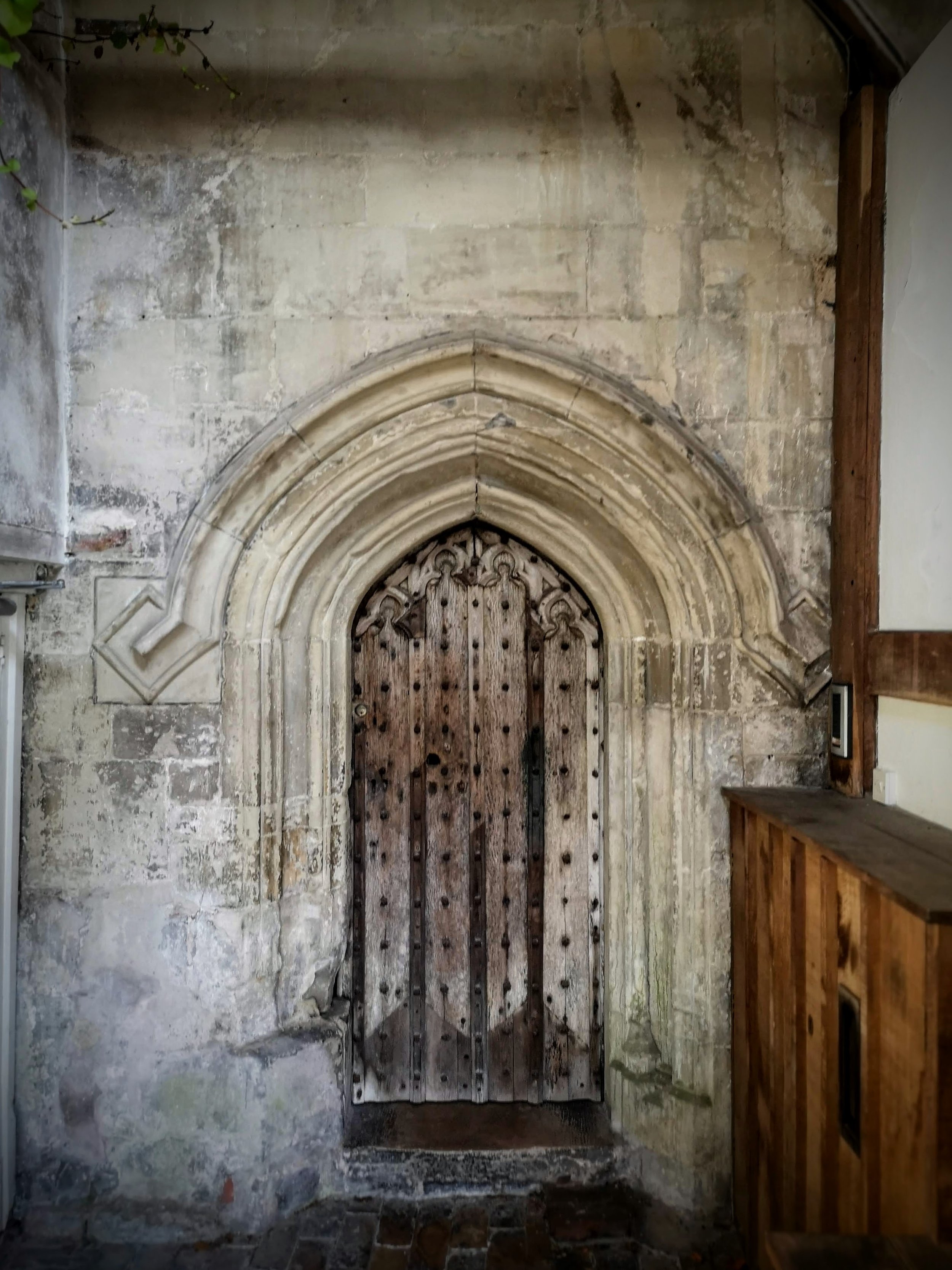 A medieval wooden door with metal studs set in an arched stone frame, attached to an aged stone wall.
