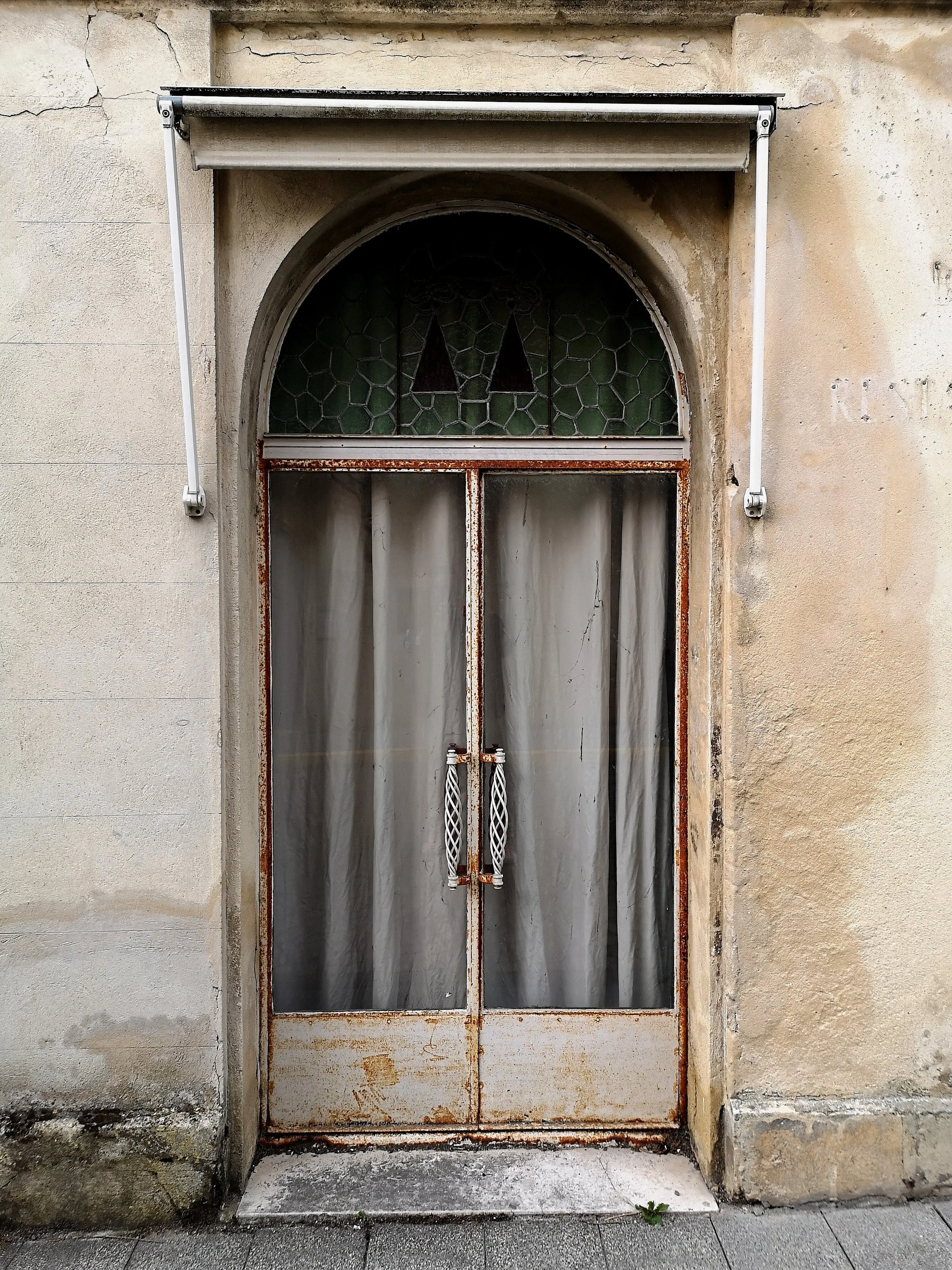 Old, rusty glass door with a curtain behind, above a small arched stained glass window, set in a weathered beige wall.