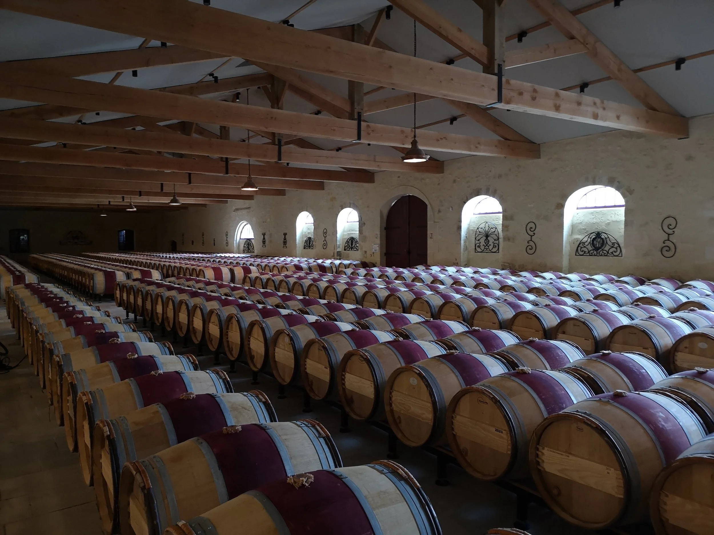 Wine barrels stored in a winery with wooden beams and arched windows.