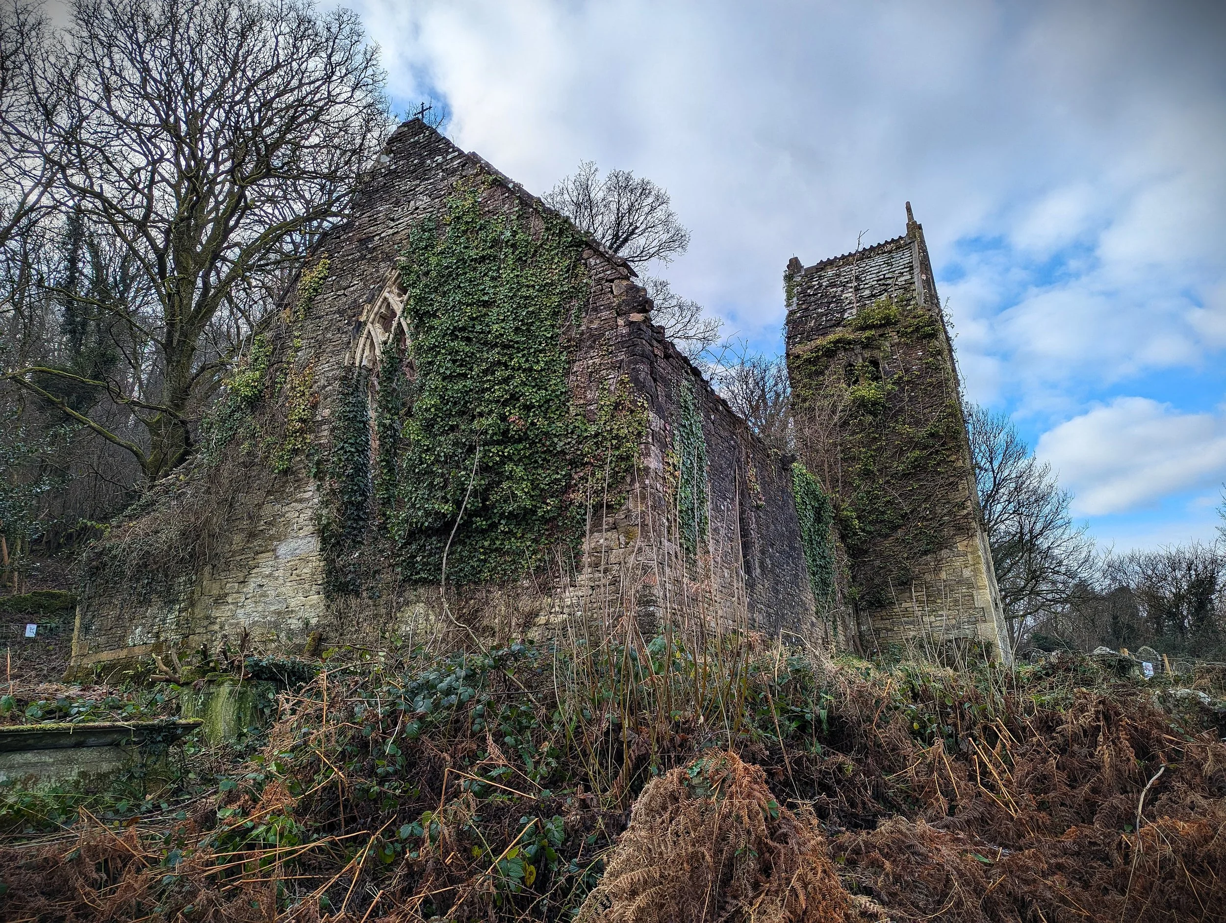 An old, abandoned stone church with a tall, leaning tower, surrounded by overgrown vegetation and leafless trees, under a partly cloudy sky.