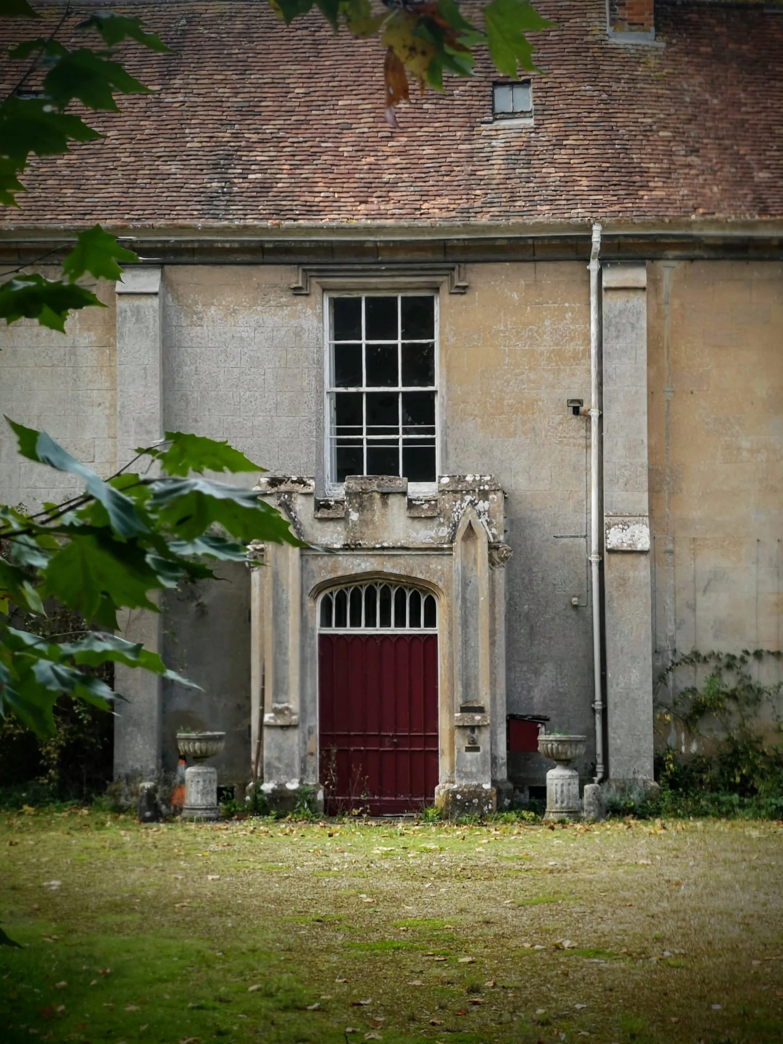 An old building with a weathered stone facade, a large arched window above a red door, and surrounded by greenery.