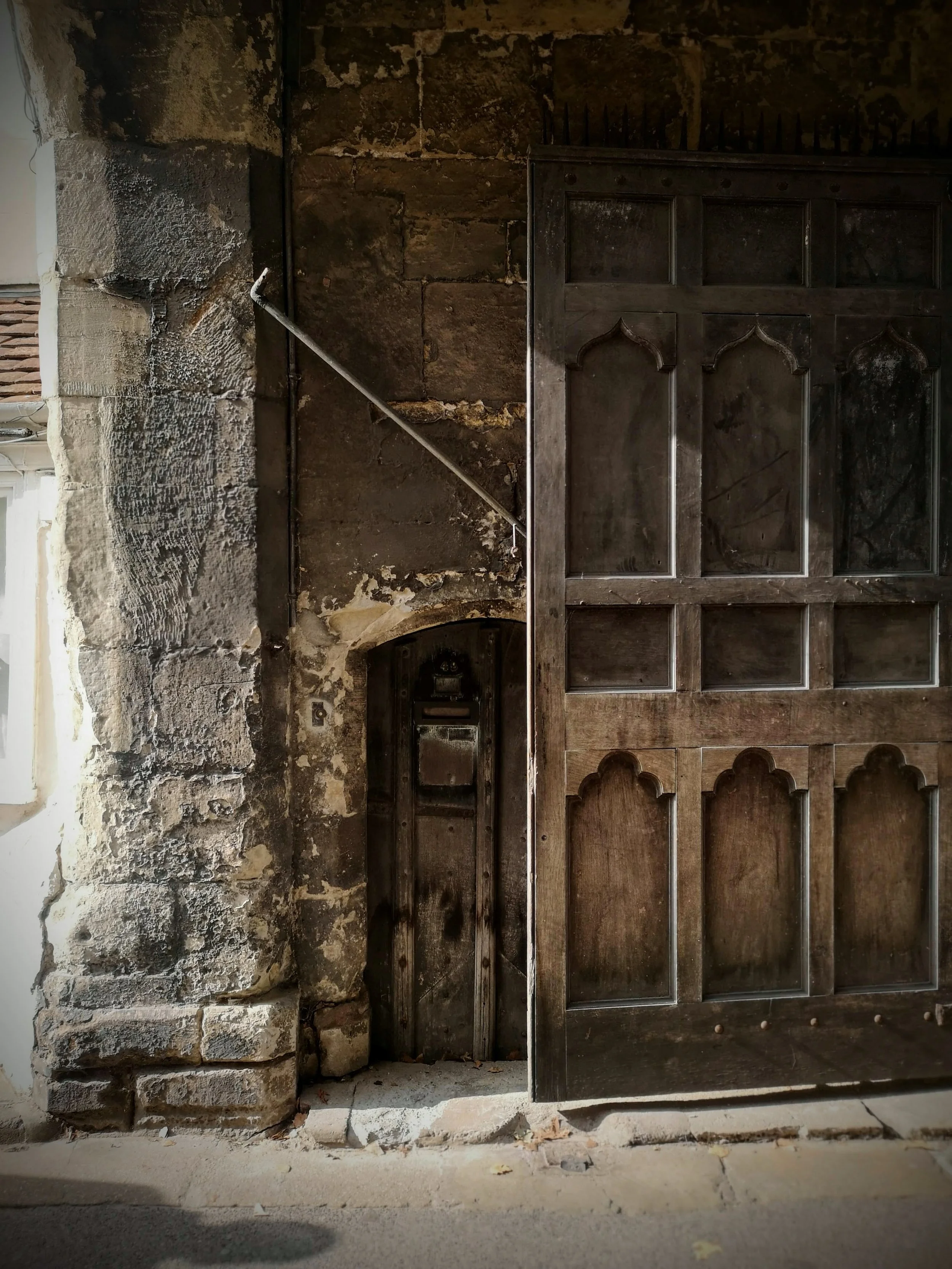 An old stone wall and a large wooden door partially opened, revealing a dark, small space behind it. The door has an ornate design with rectangular and arched panels.