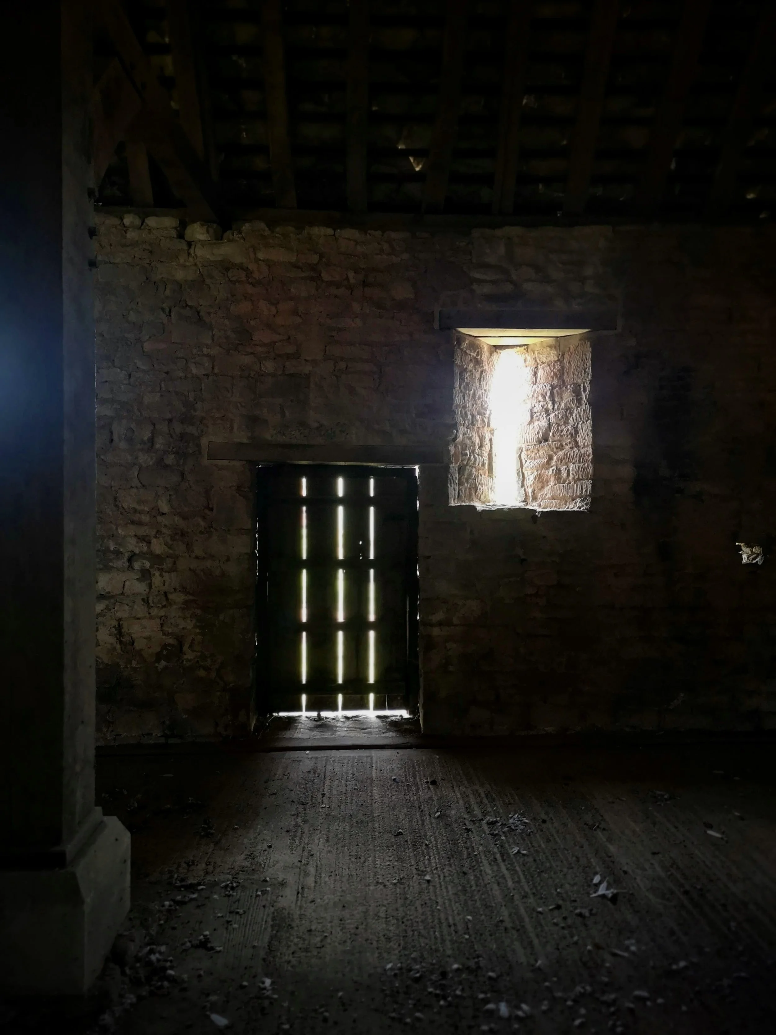 Dark interior of a rustic stone building with a small window and a wooden door, both letting in light.