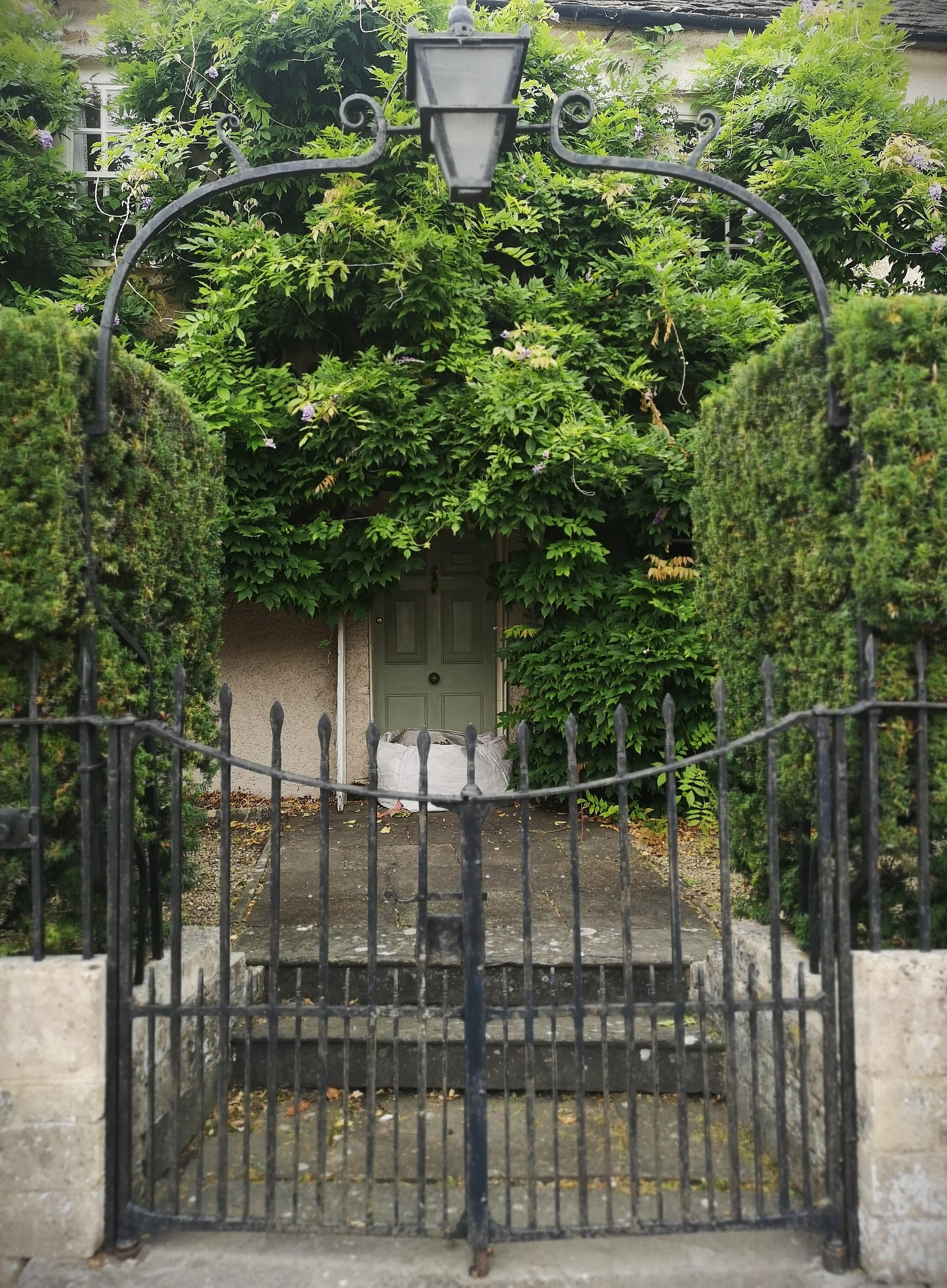 A black iron gate with a decorative arch and a hanging lantern, opening to a small stone pathway leading to a front door surrounded by lush green bushes and climbing plants.