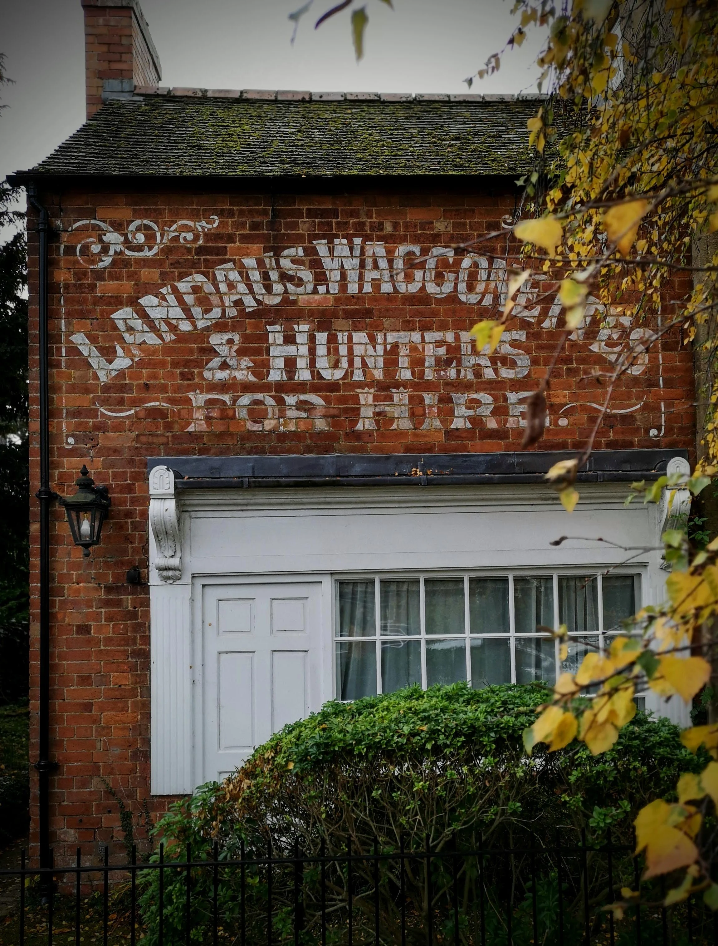 A brick building with a painted sign that reads "LAVIDAS WAGON & HUNTERS FOR HIRE." The sign is faded. There is a white framed window with a hedge in front and a black lamp on the left side of the building.