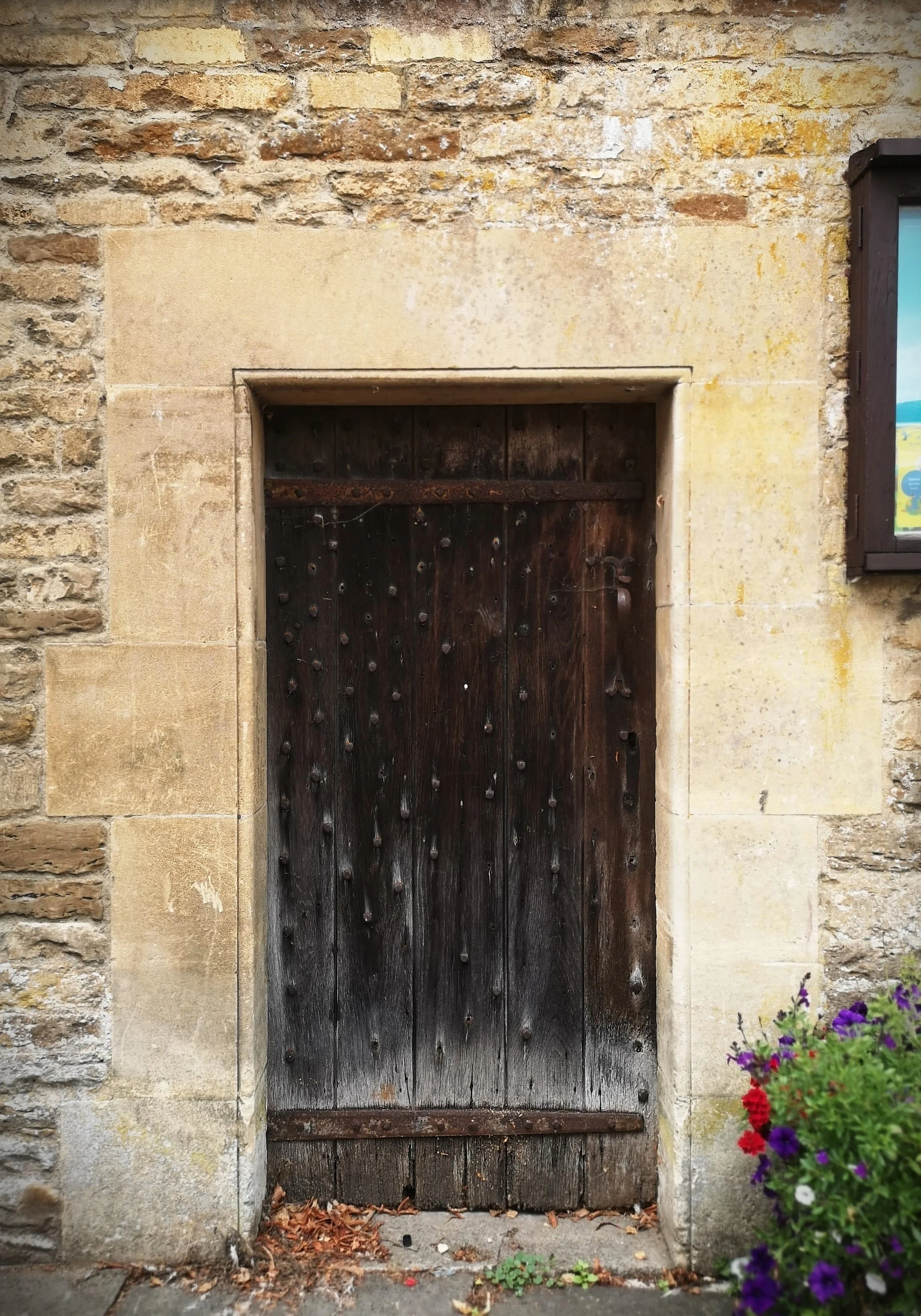 Old wooden door with rusted metal hinges and nails, set in a stone wall with a flowerpot of purple, red, and white flowers to the right.