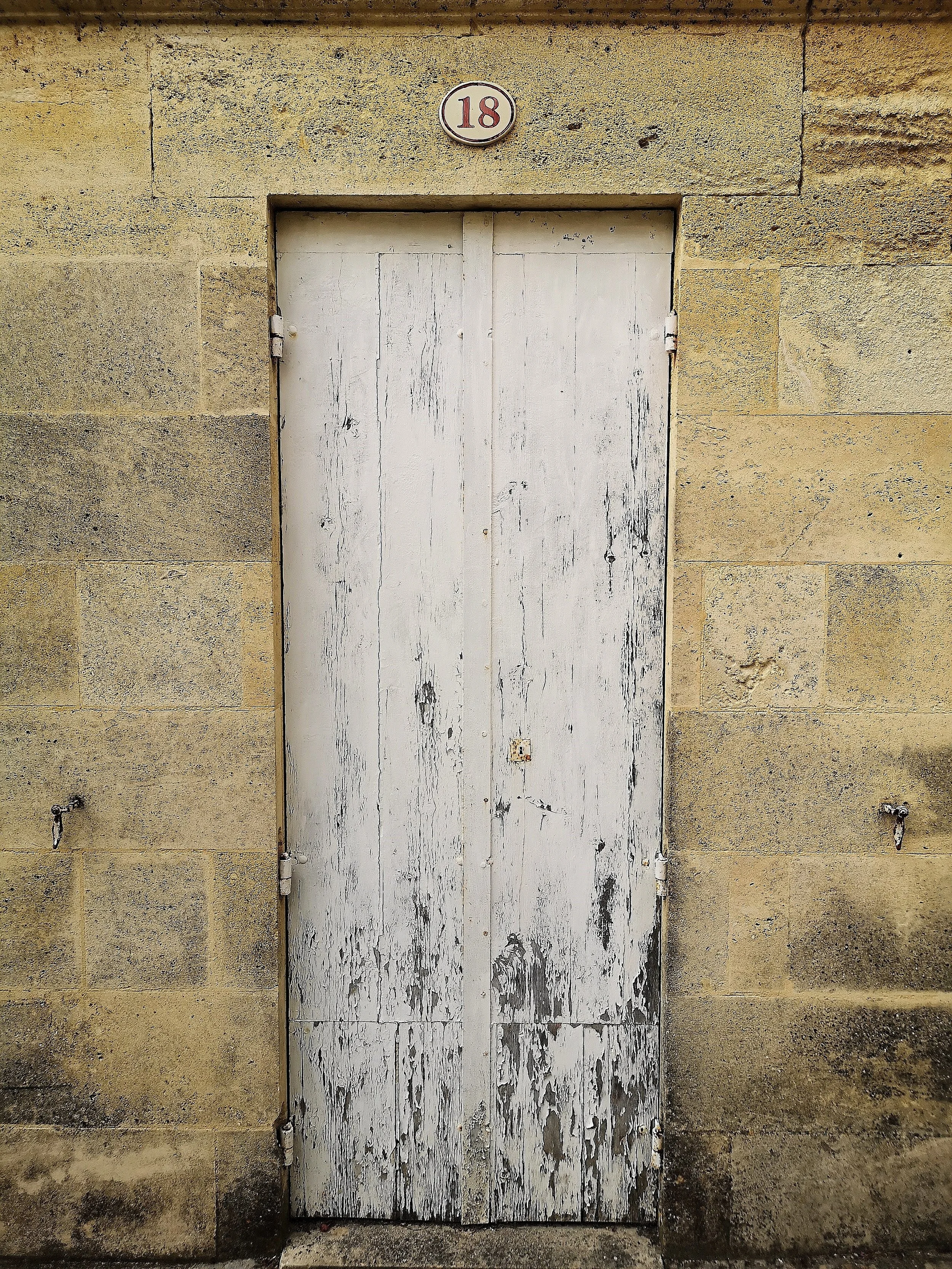 Weathered white wooden door set in a yellow brick wall with a small oval sign displaying the number 18 above it.