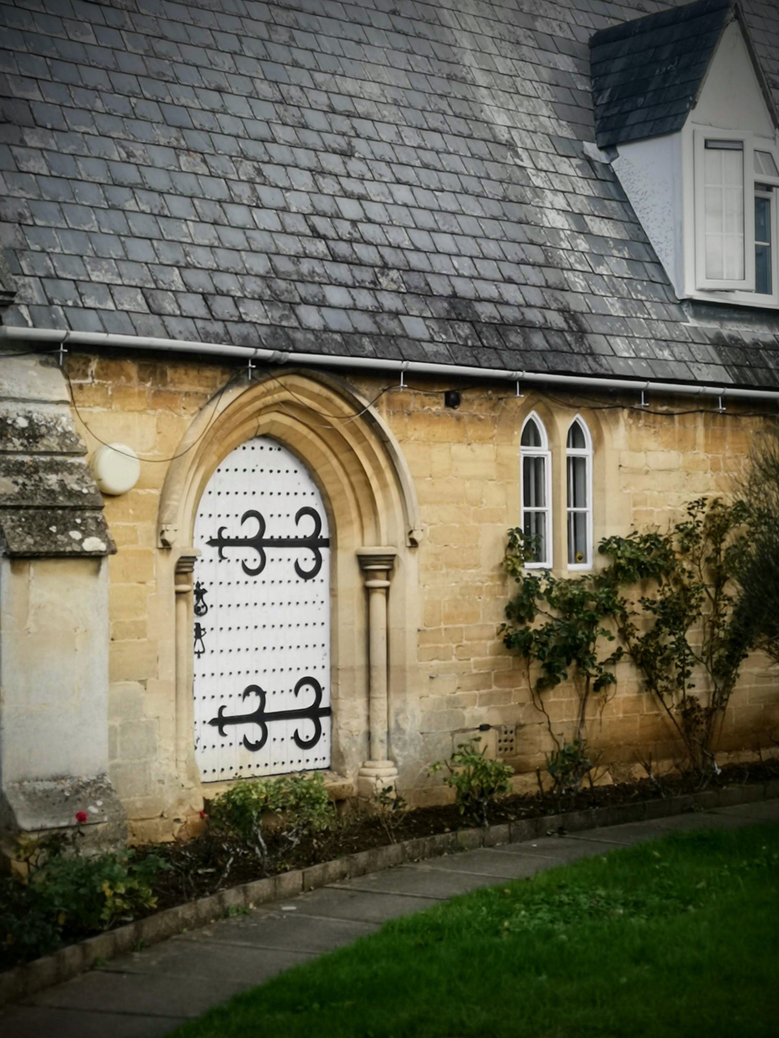 A stone building with a arched white door decorated with black wrought iron designs, small windows with pointed arches, and a grey shingled roof.