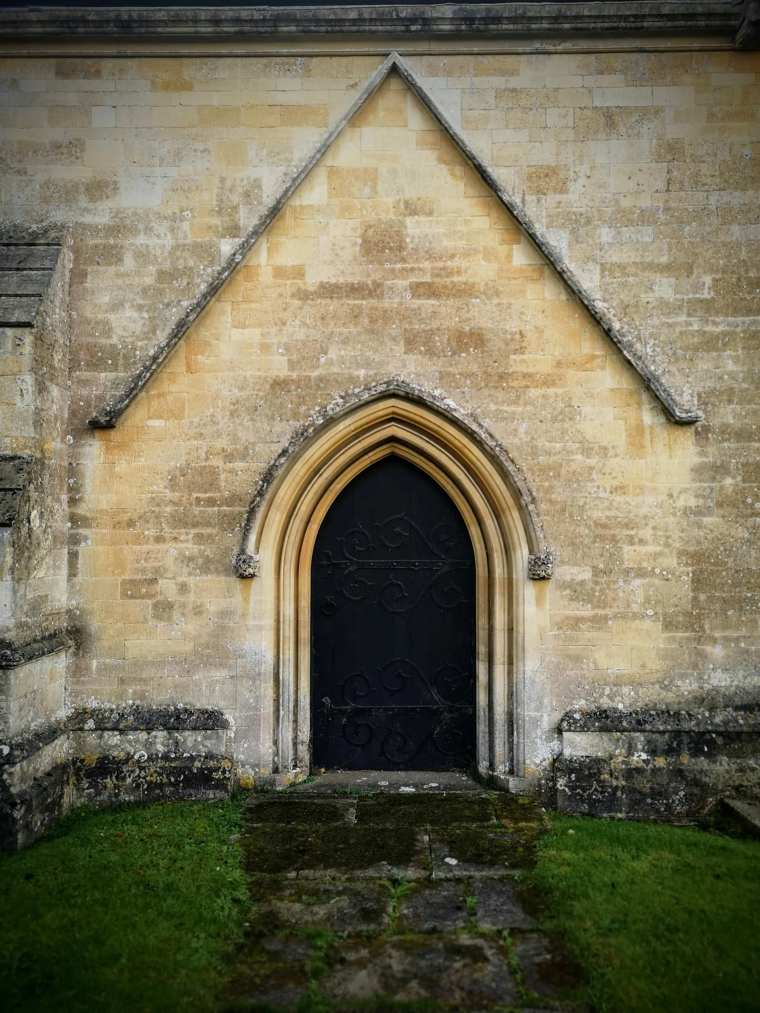 A small, arched black door with decorative ironwork is set into a beige stone wall of an old church or historic building. There is moss and grass on the ground in front of the door.