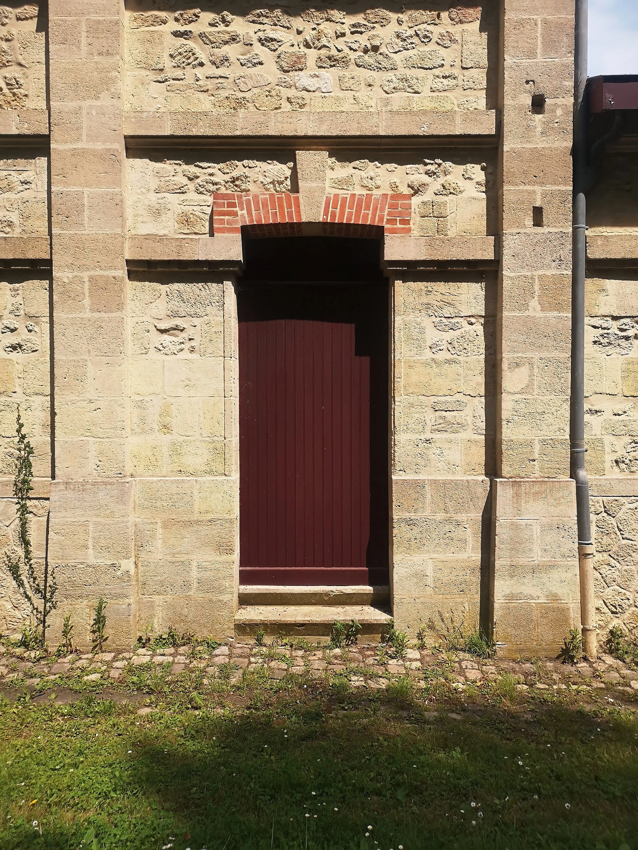 A tall, narrow, maroon wooden door set into a stone building, with a small, three-step concrete porch in front. The building has a mix of beige and light brown stones with some red brick accents above the door. There are some small plants and grass a