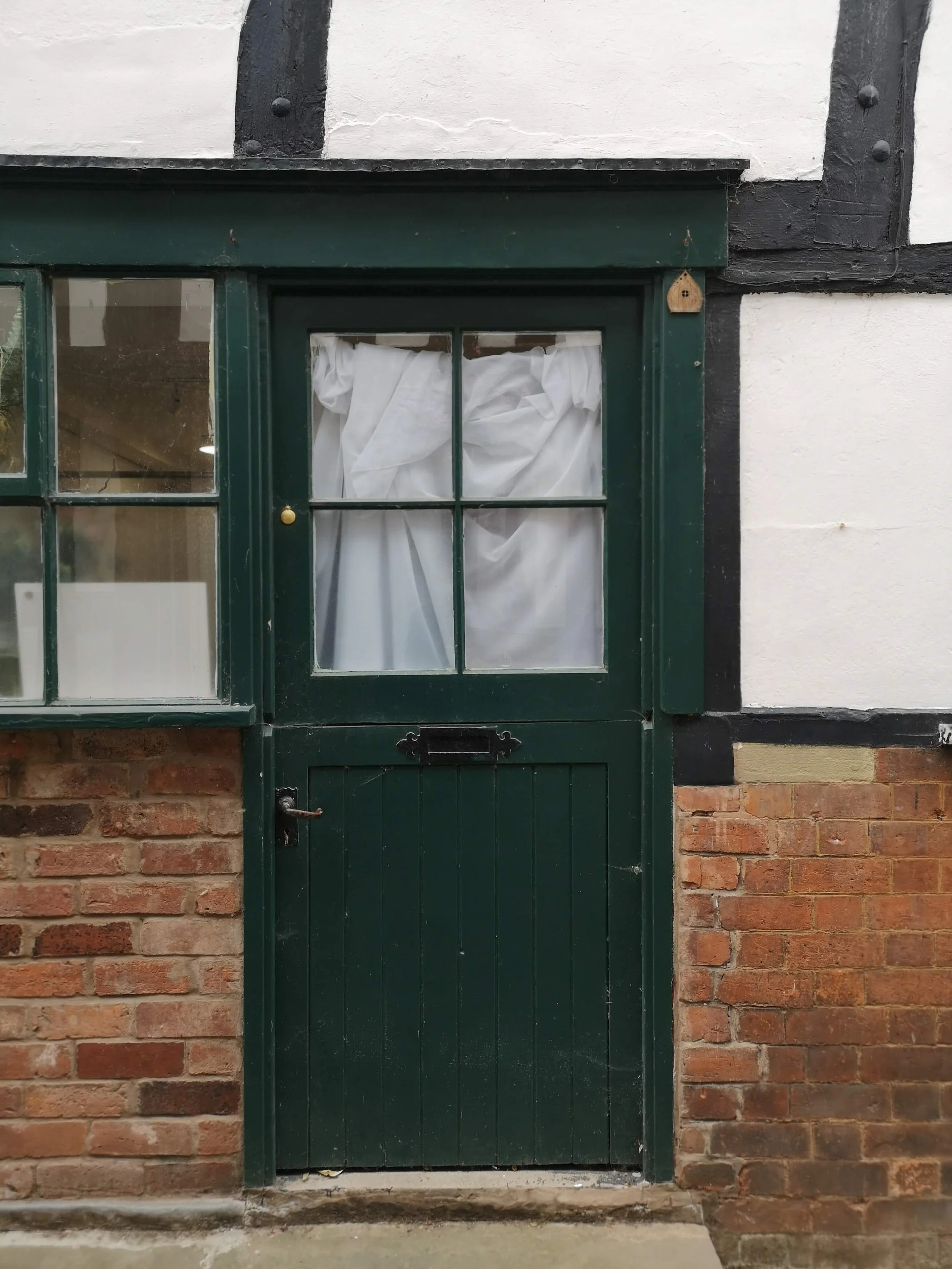 Green wooden door with window panes, partly covered by white curtains, set in a brick and white plaster wall.