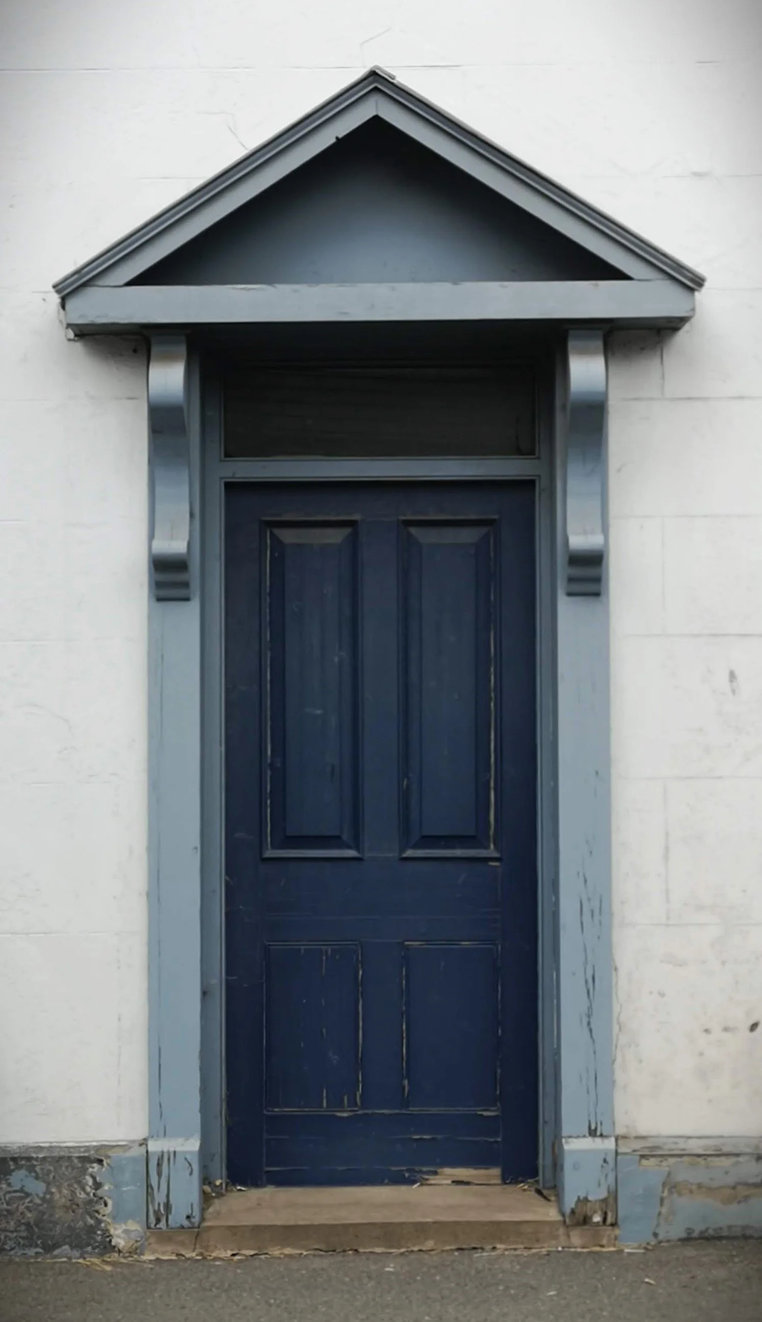 A dark blue wooden door with a rectangular window above, set in a white brick wall. The door has decorative panels and a small wooden step at the bottom. A triangular gray porch roof with metallic brackets extends above the door.