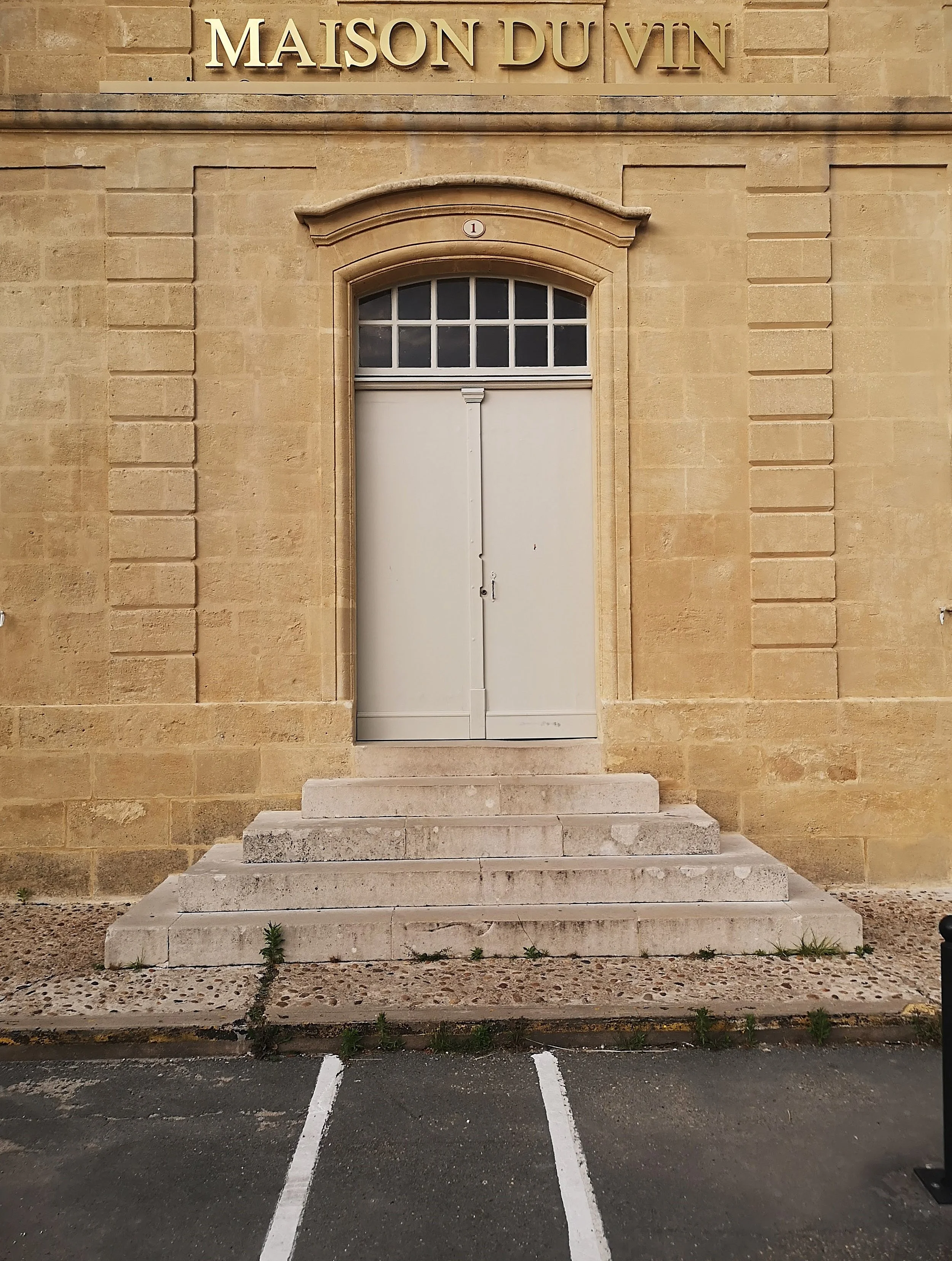 Front view of a beige stone building with a sign reading 'MAISON DUVIN' above a set of three stone steps leading to a closed white door. There is a small window with multiple panes above the door. Parking spaces with white lines are in front of the s