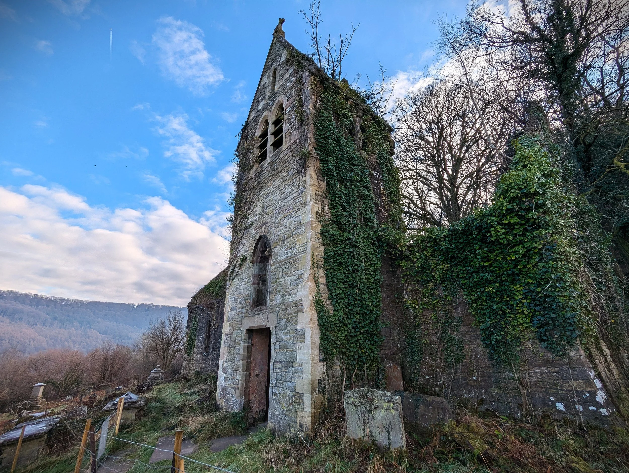 Old stone church building with ivy growing on its walls, set against a partly cloudy sky and trees in the background.