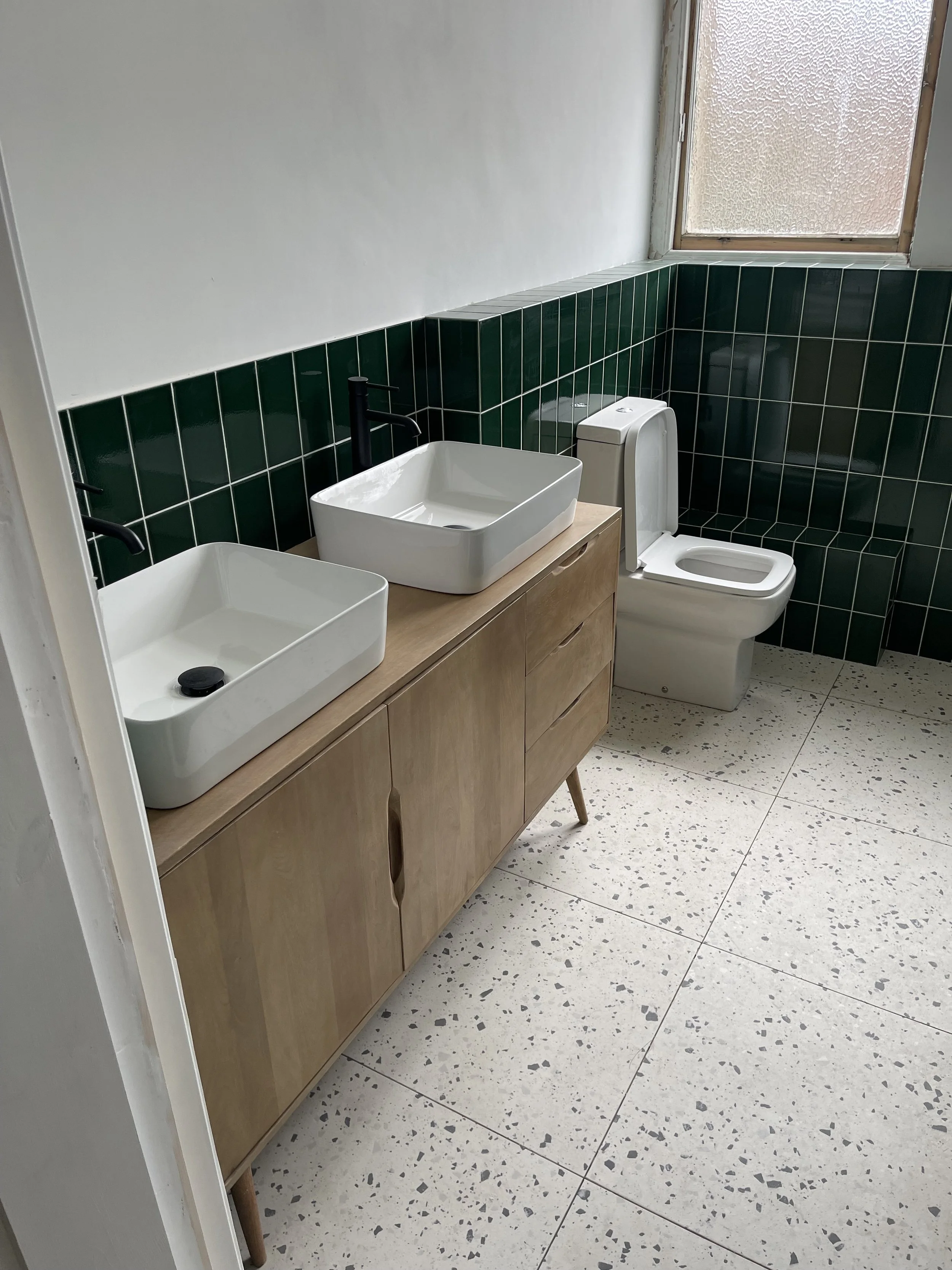 A modern bathroom featuring white square sinks with black faucets set on a light wood vanity, a white toilet, green tiled wall with black grout, a window with textured glass, and terrazzo-style floor tiles.