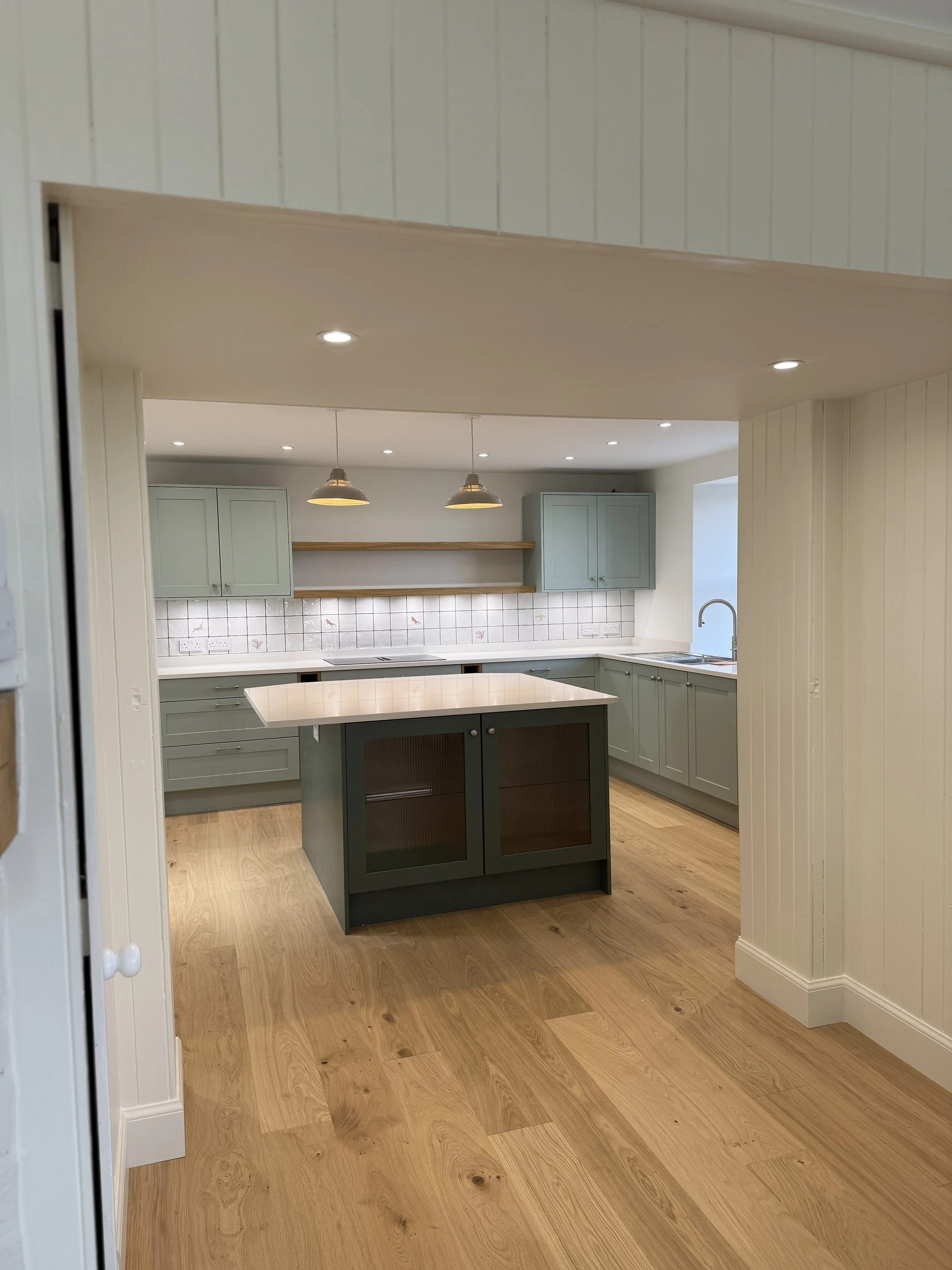 Photo of a modern kitchen with light green cabinets, a white countertop, a kitchen island with dark green doors, wooden flooring, and pendant lights hanging from the ceiling.