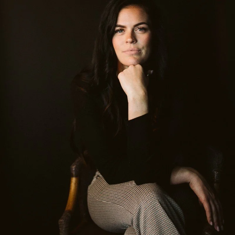 Woman sitting with chin resting on hand, wearing a black top and checkered pants, against a dark background.