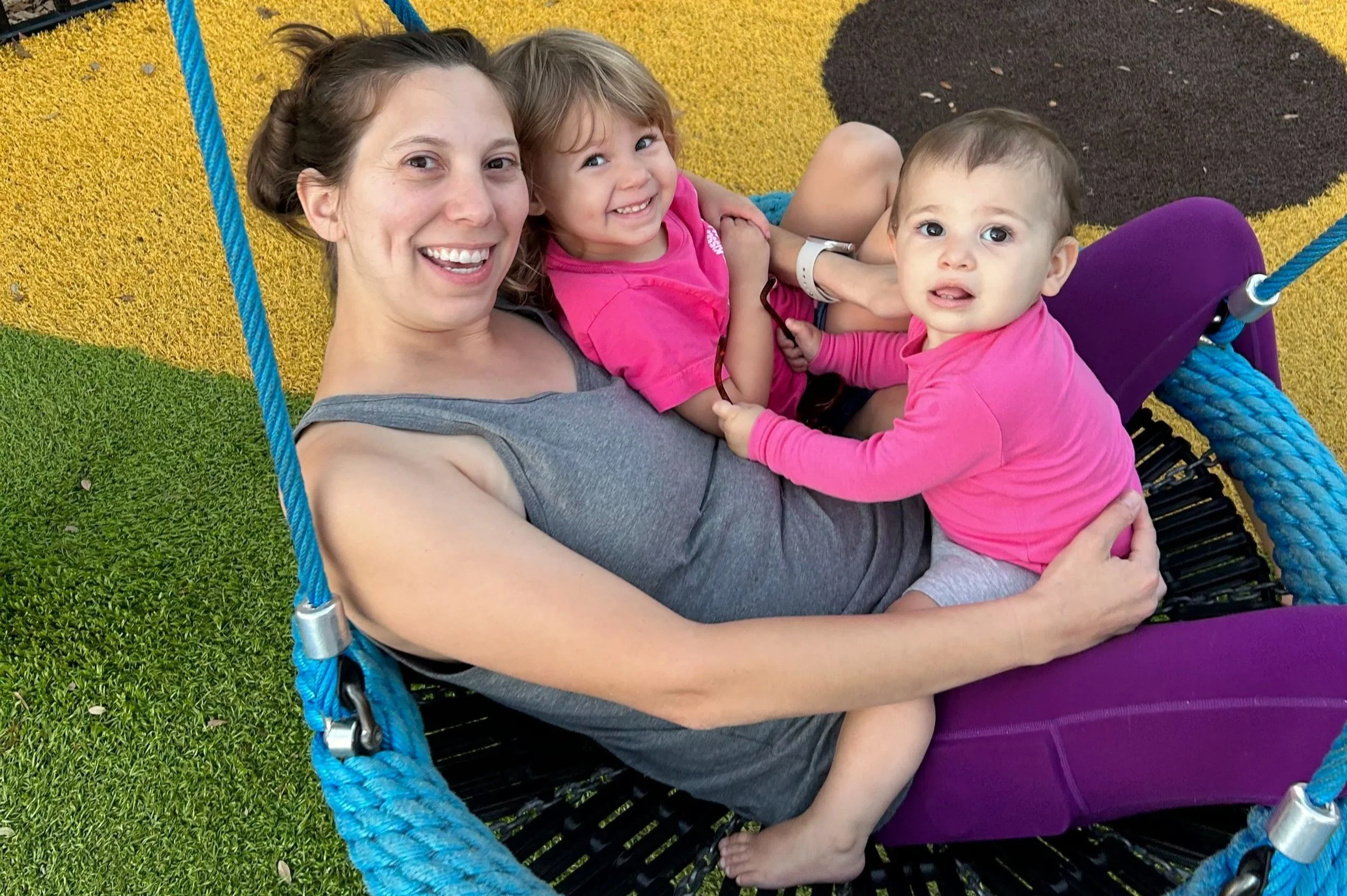 A person with two children sitting in a circular swing on a playground. The person is wearing a gray tank top and purple pants, while the children are wearing pink shirts. The ground is covered with colorful turf.