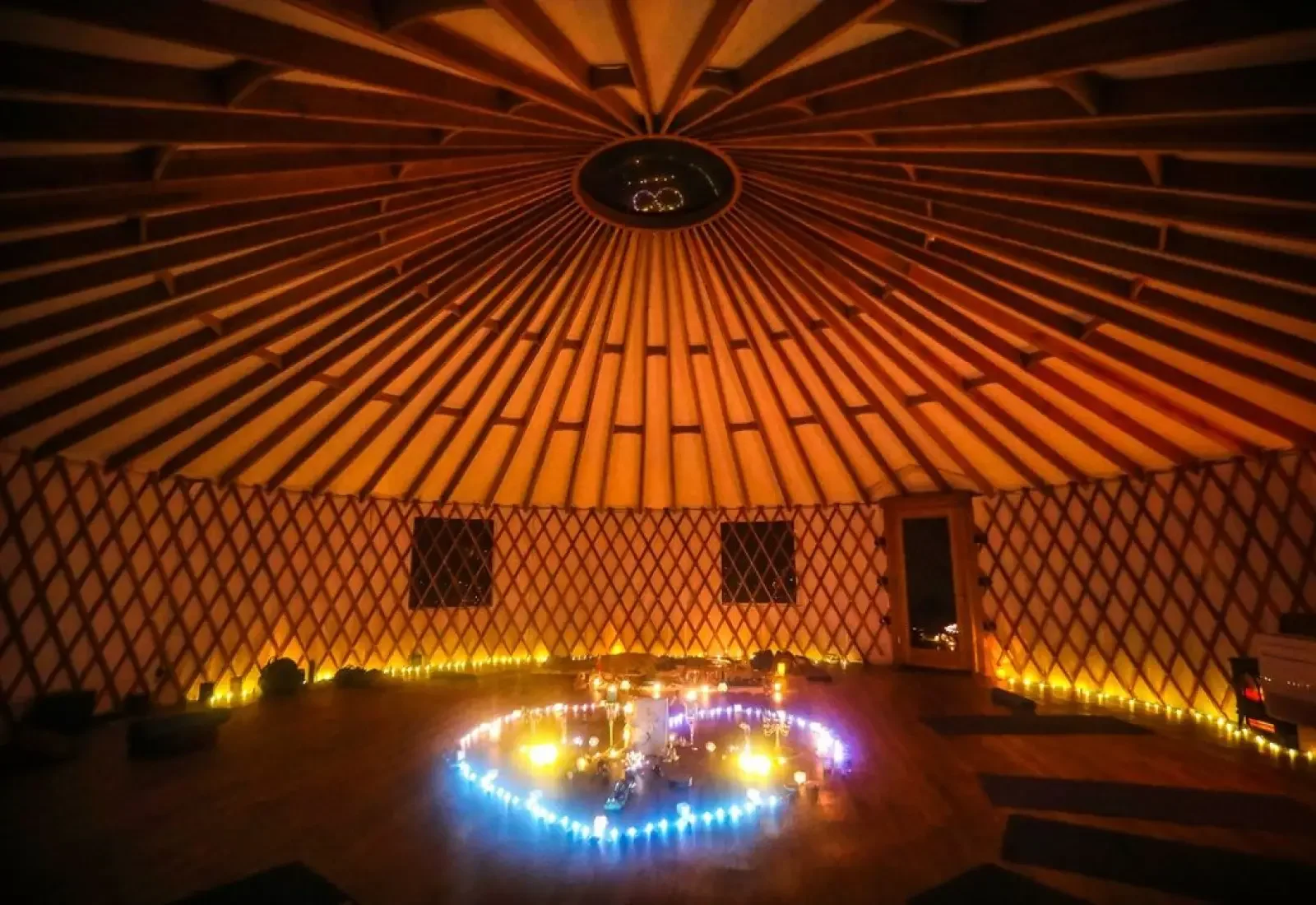 Interior view of a round yurt with a wooden lattice wall and a conical wooden ceiling, illuminated with warm string lights and candles in a heart-shaped arrangement on the floor.