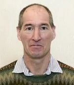 Photo of a man with a light complexion, short brown hair, and wearing a patterned sweater over a collared shirt, standing in front of a plain light-colored wall.