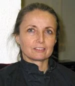 A woman with brown hair tied back, wearing a black top, standing indoors.