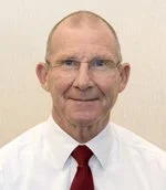 A smiling elderly man with glasses, short hair, wearing a white shirt and red tie.